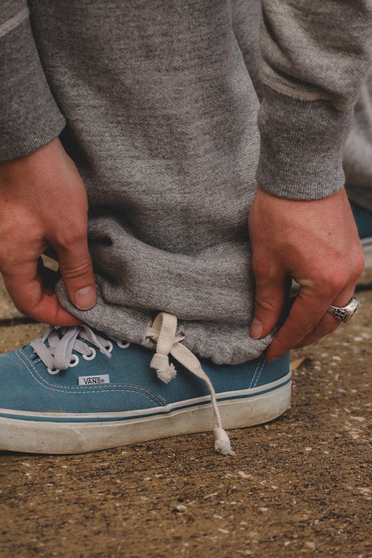 A close-up of a person's hands pulling down the hem of gray sweatpants to reveal a blue Vans sneaker underneath, set against a textured concrete ground.