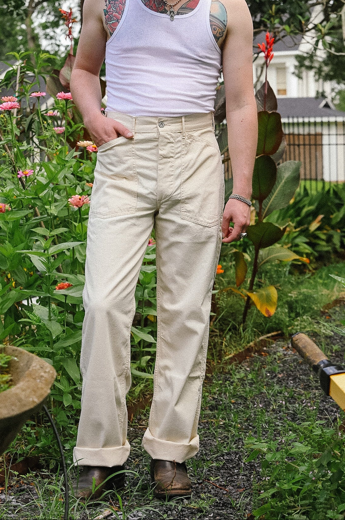 Man standing in a garden wearing a white tank top and beige pants.
