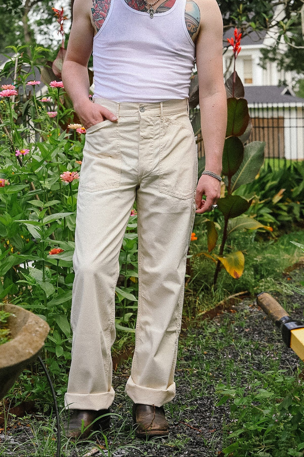 Man standing in a garden wearing a white tank top and beige pants.