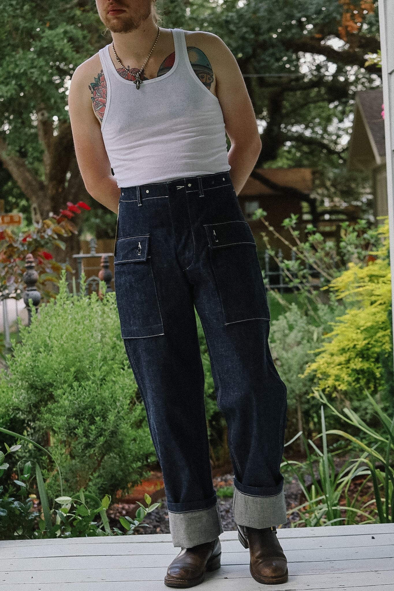 Person wearing a white tank top and dark jeans standing in a garden.