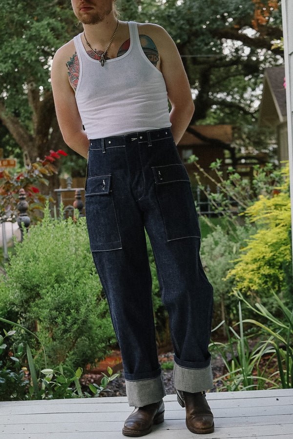 Person wearing a white tank top and dark jeans standing in a garden.