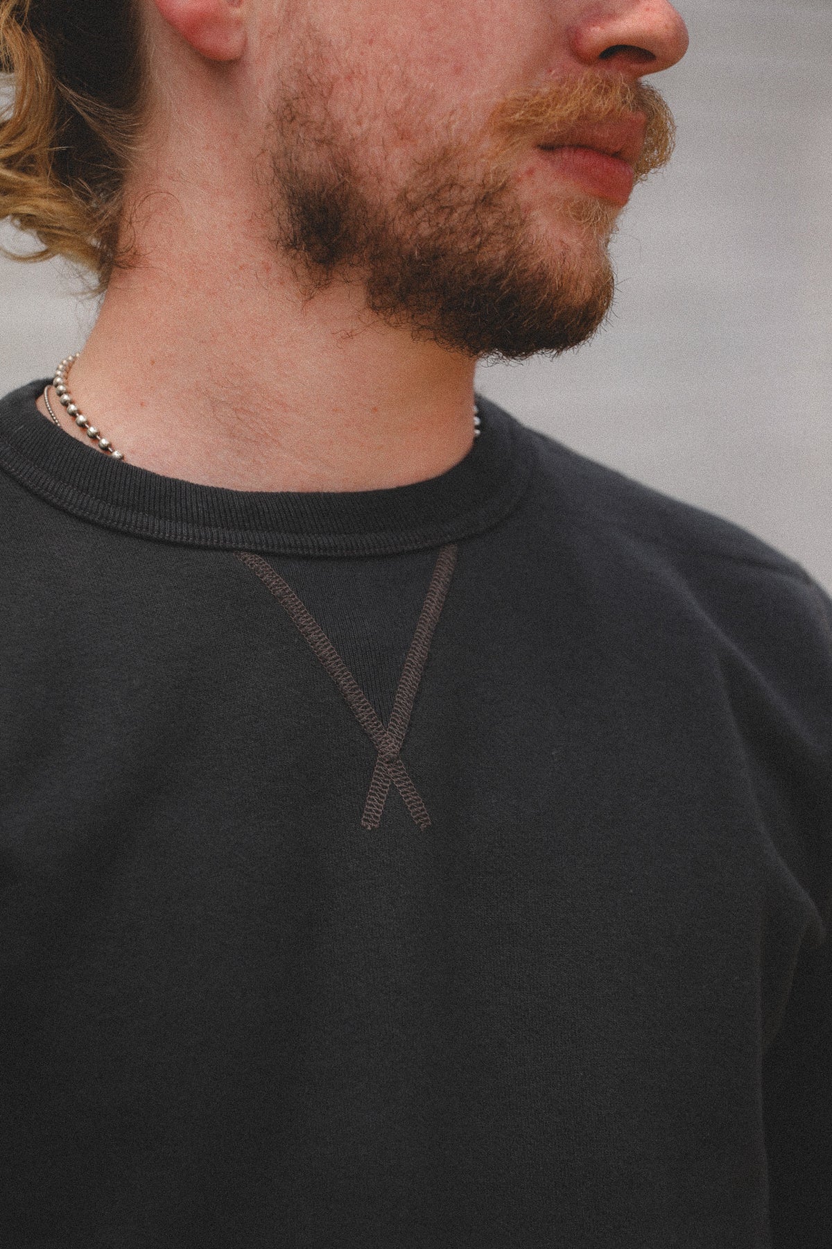 Close-up of a man wearing a dark charcoal crewneck sweatshirt with a subtle tonal embroidered 'Y' logo on the chest, paired with a silver beaded necklace.