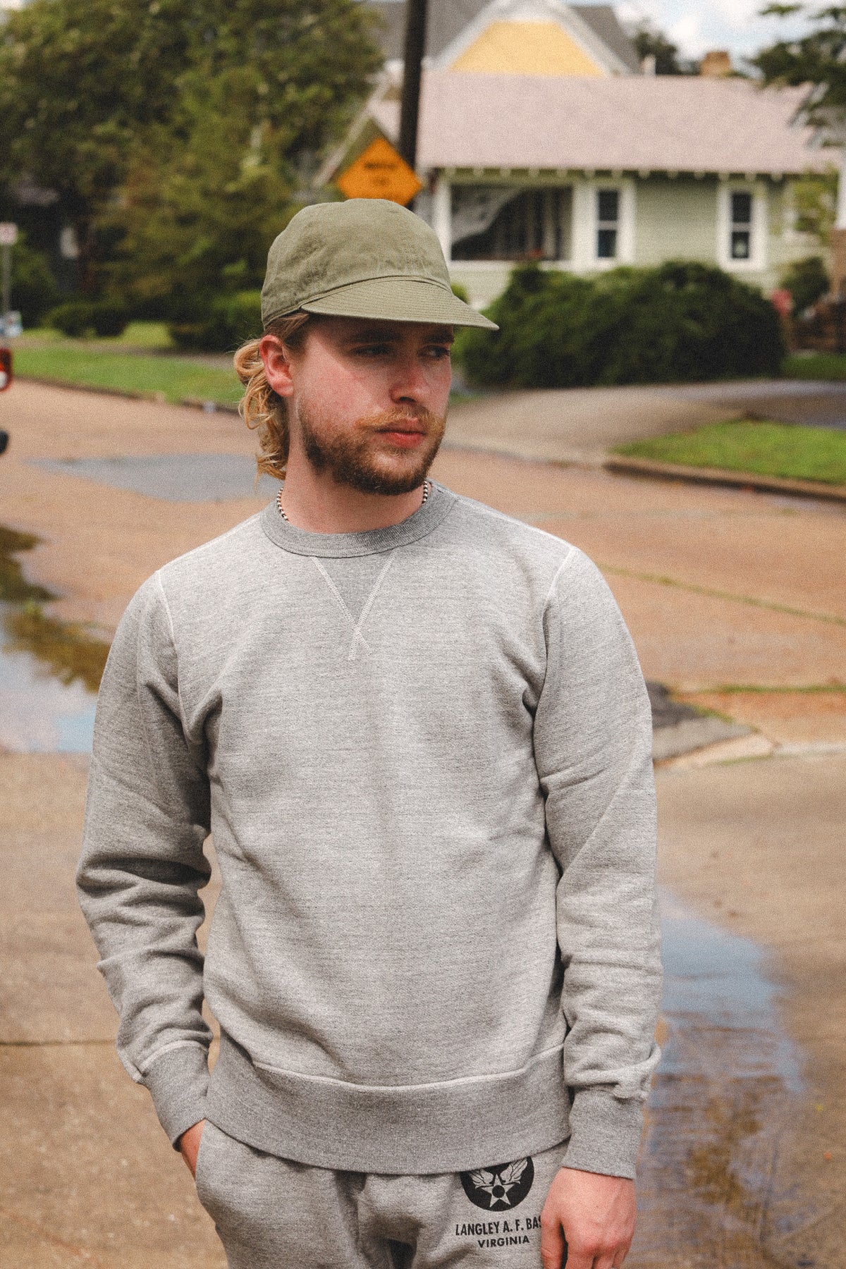 A man wearing a light gray crewneck sweatshirt and matching sweatpants stands on a wet residential street, wearing an olive green baseball cap. The sweatshirt features a small white embroidered cross on the chest.