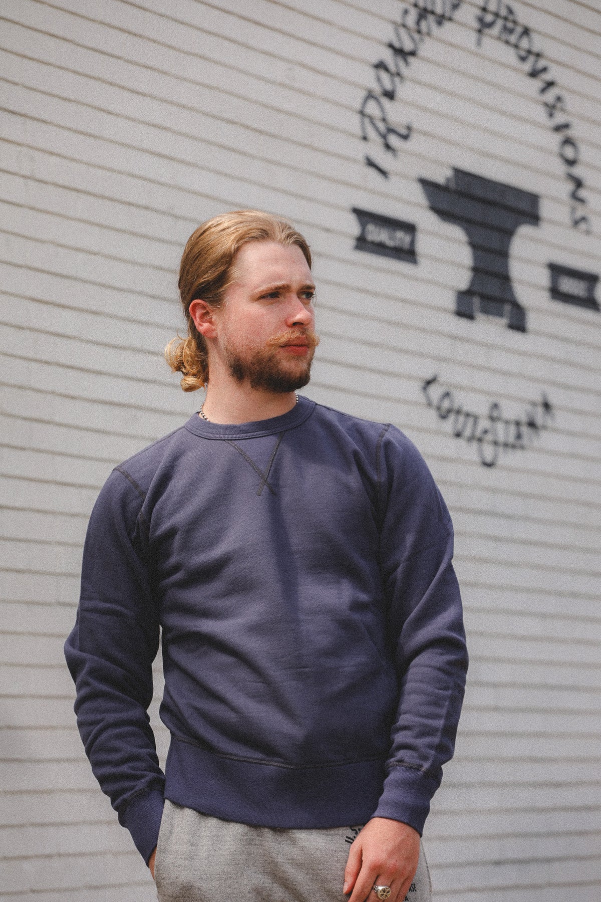 A man with blonde hair tied back and a beard, wearing a navy blue crewneck sweatshirt and gray sweatpants, standing in front of a white brick wall with a black anvil graphic and the words 'Iron Forge Provisions' and 'Koldhane'.
