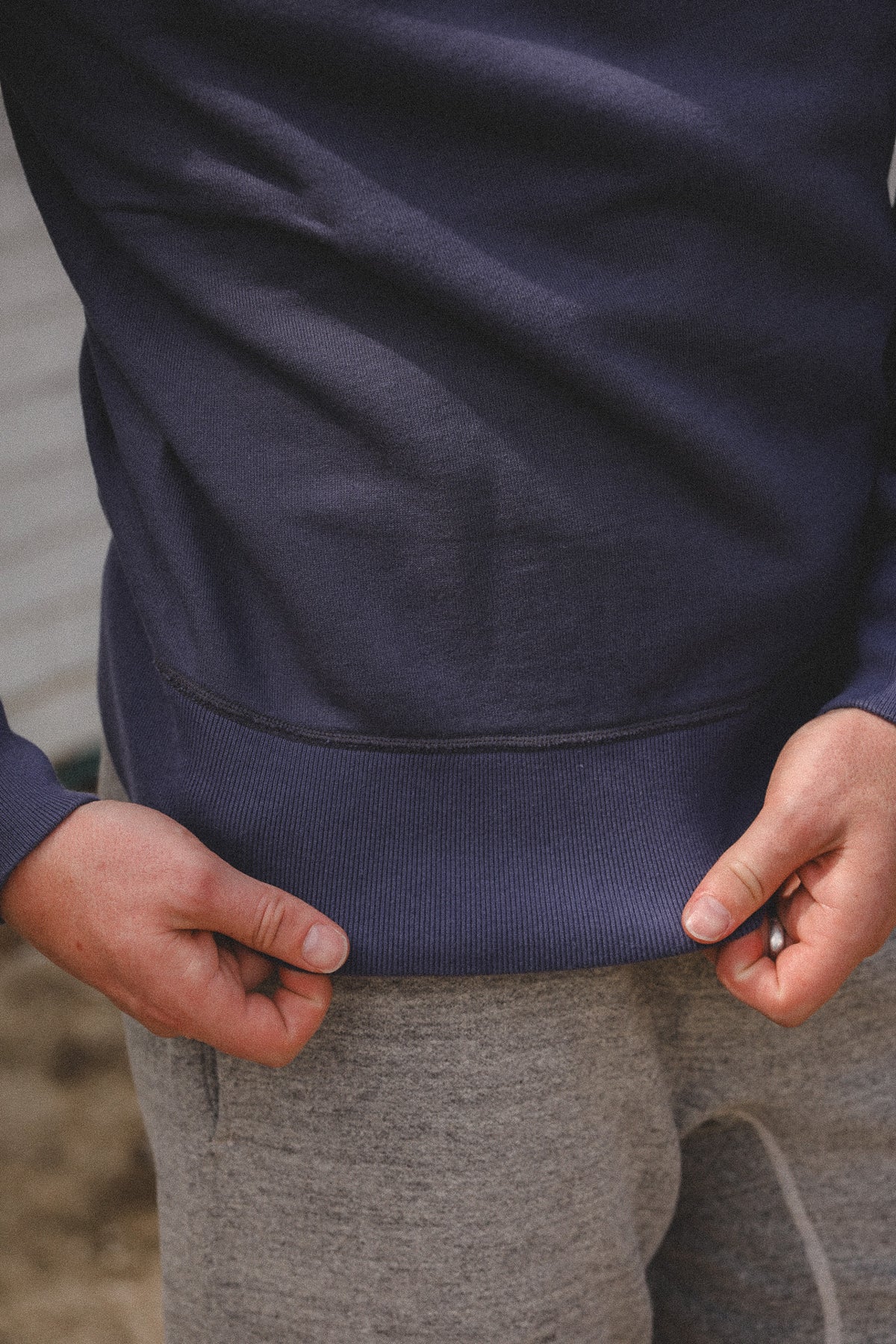 Close-up of a person wearing a navy blue ribbed sweatshirt and gray sweatpants, pulling the hem of the navy sweatshirt outward with both hands.