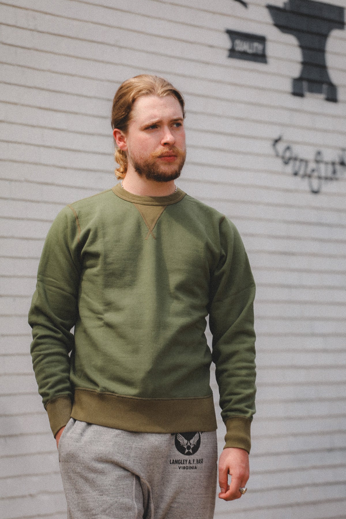 A man with blonde hair in a side braid wearing an olive green crewneck sweatshirt and gray sweatpants, standing against a white brick wall with black graffiti.