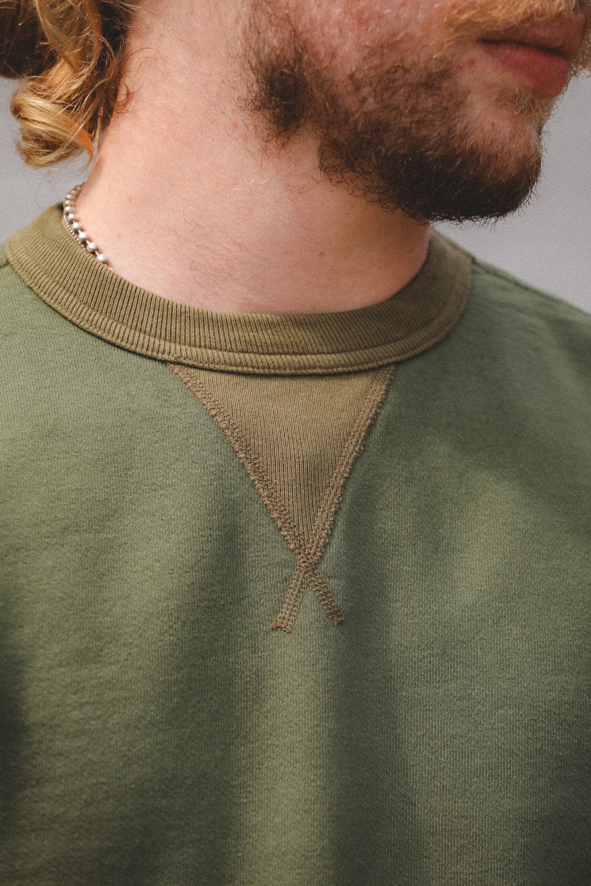 Close-up of a person wearing an olive green crewneck sweatshirt with a contrasting ribbed collar and a small triangular chest detail.