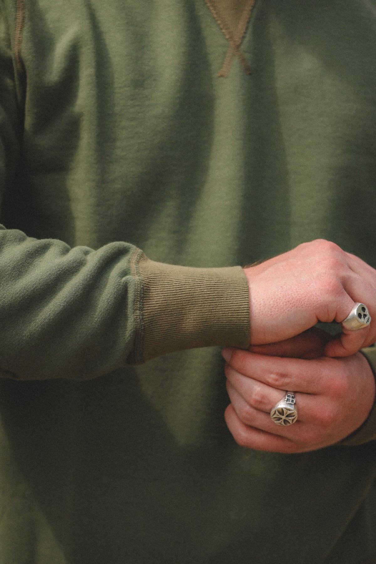 Close-up of a person wearing an olive green crewneck sweatshirt, showing the ribbed cuffs and hands with silver rings.