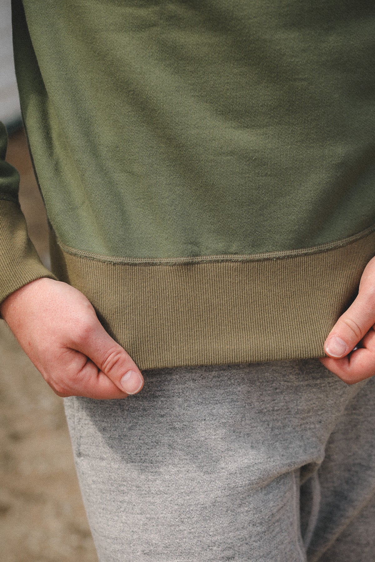 Close-up of a person wearing an olive green long-sleeve top with a ribbed hem, paired with gray sweatpants, with hands pulling the hem slightly to show the fabric stretch.