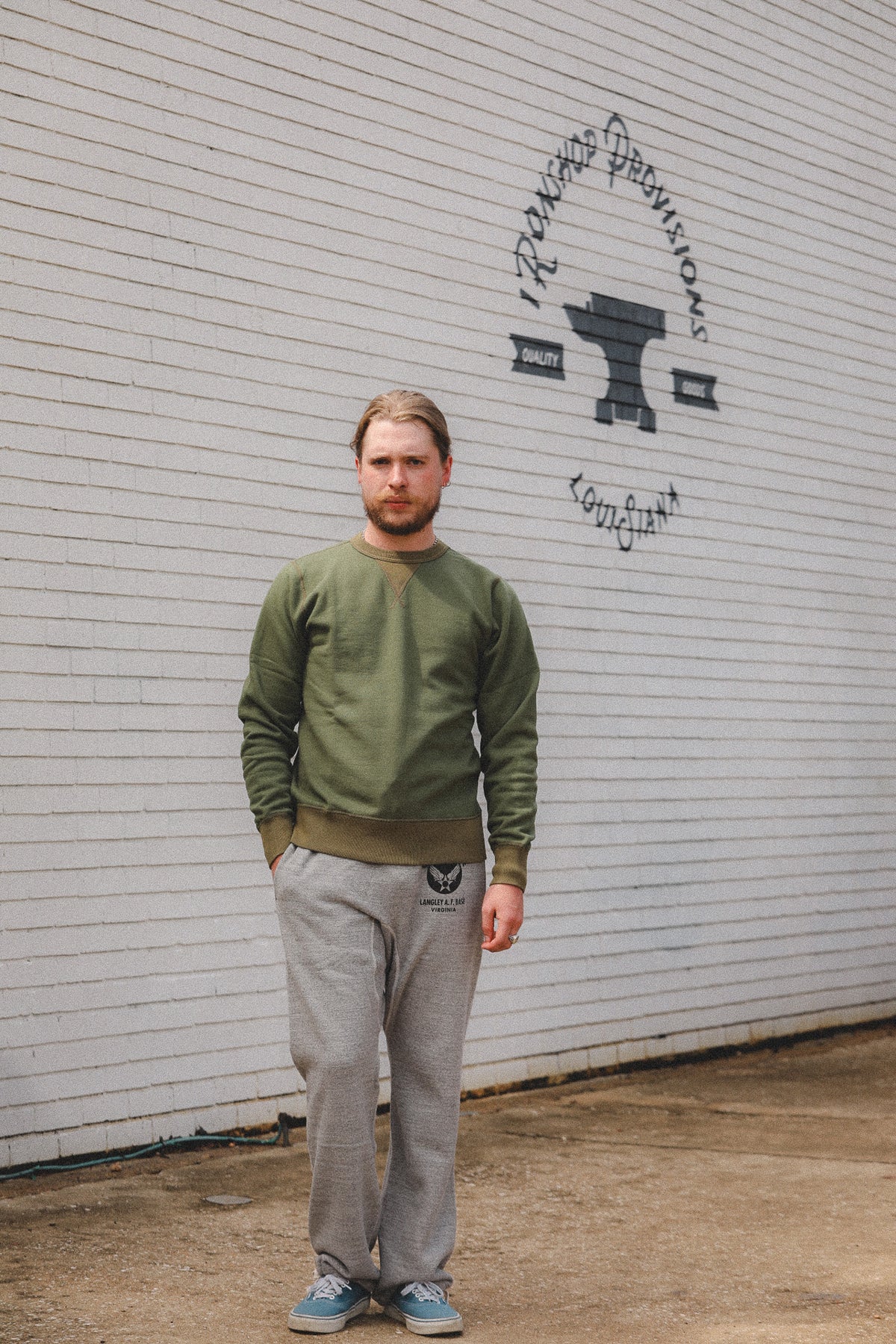A man wearing an olive green crewneck sweatshirt and gray sweatpants stands in front of a white brick wall with a black anvil logo and the words 'Forging Possibilities' and 'England'.