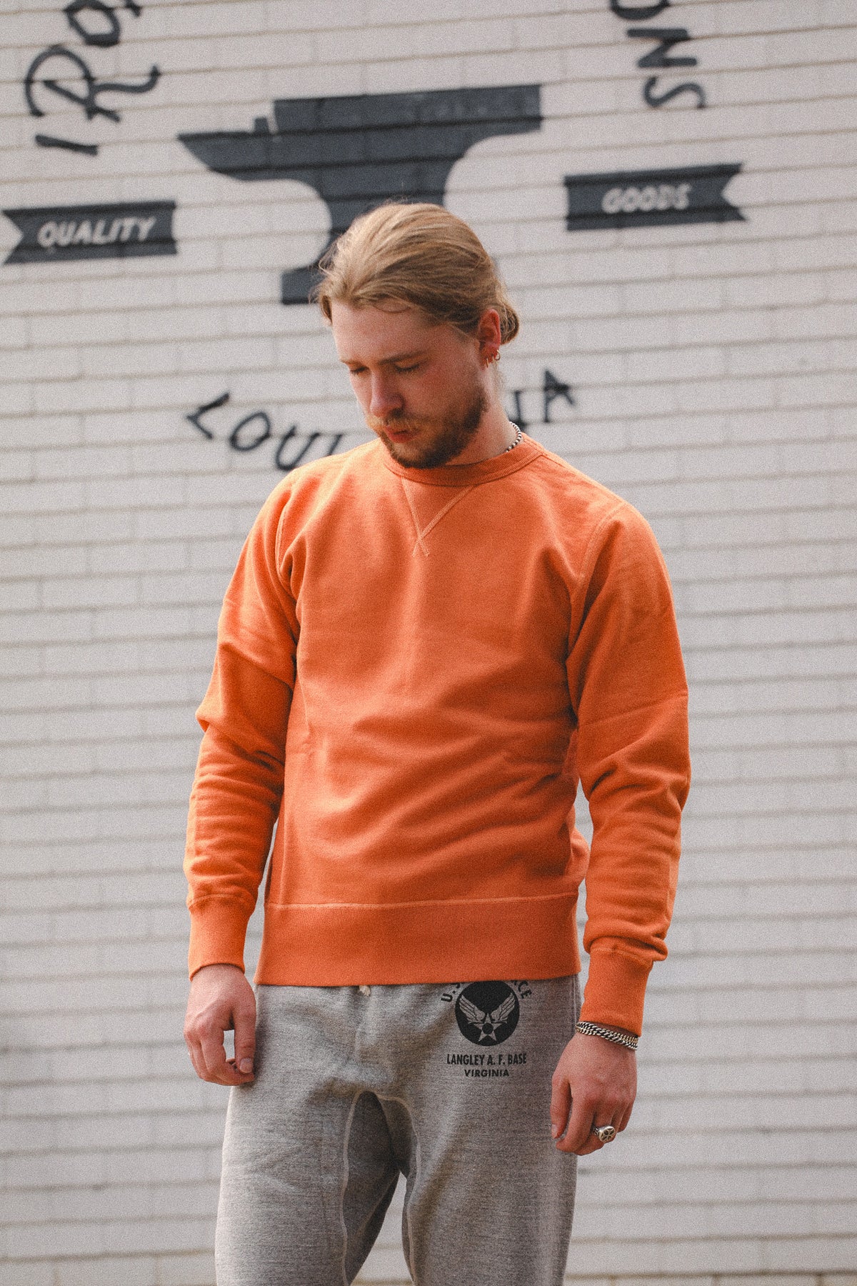 A man wearing a bright orange crewneck sweatshirt and gray sweatpants, standing in front of a white brick wall with black graphic text and an anvil design.