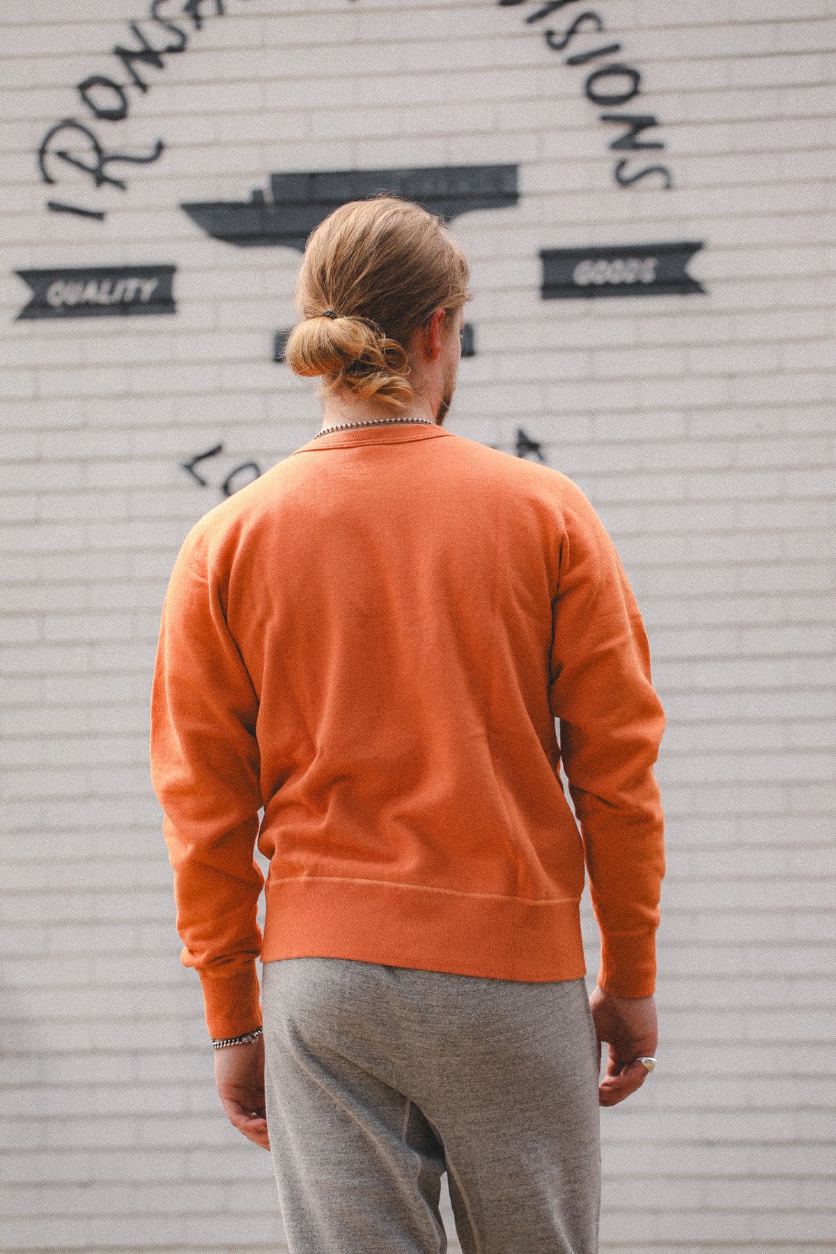 A man viewed from behind wearing a bright orange crewneck sweatshirt and gray sweatpants, standing in front of a white brick wall with black lettering.