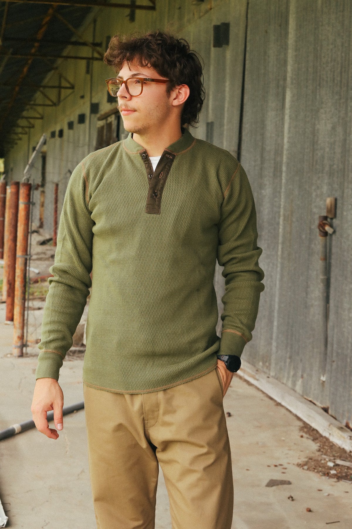 A man wearing a sage green long-sleeve henley pullover sweater with a dark brown collar detail and khaki pants, standing in an industrial corridor.