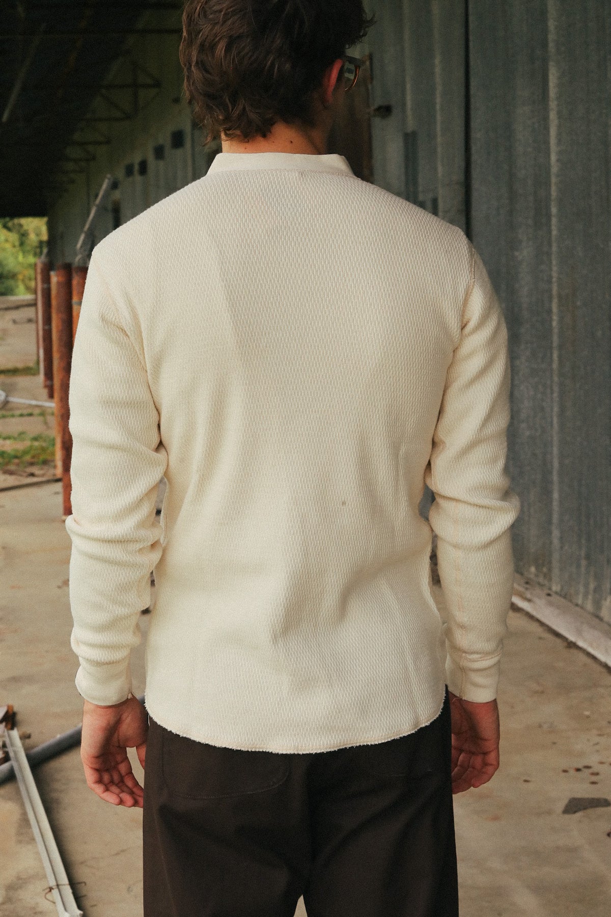 A man viewed from behind wearing a cream-colored ribbed knit long-sleeve shirt and dark brown pants, standing in an industrial setting with a corrugated metal wall.