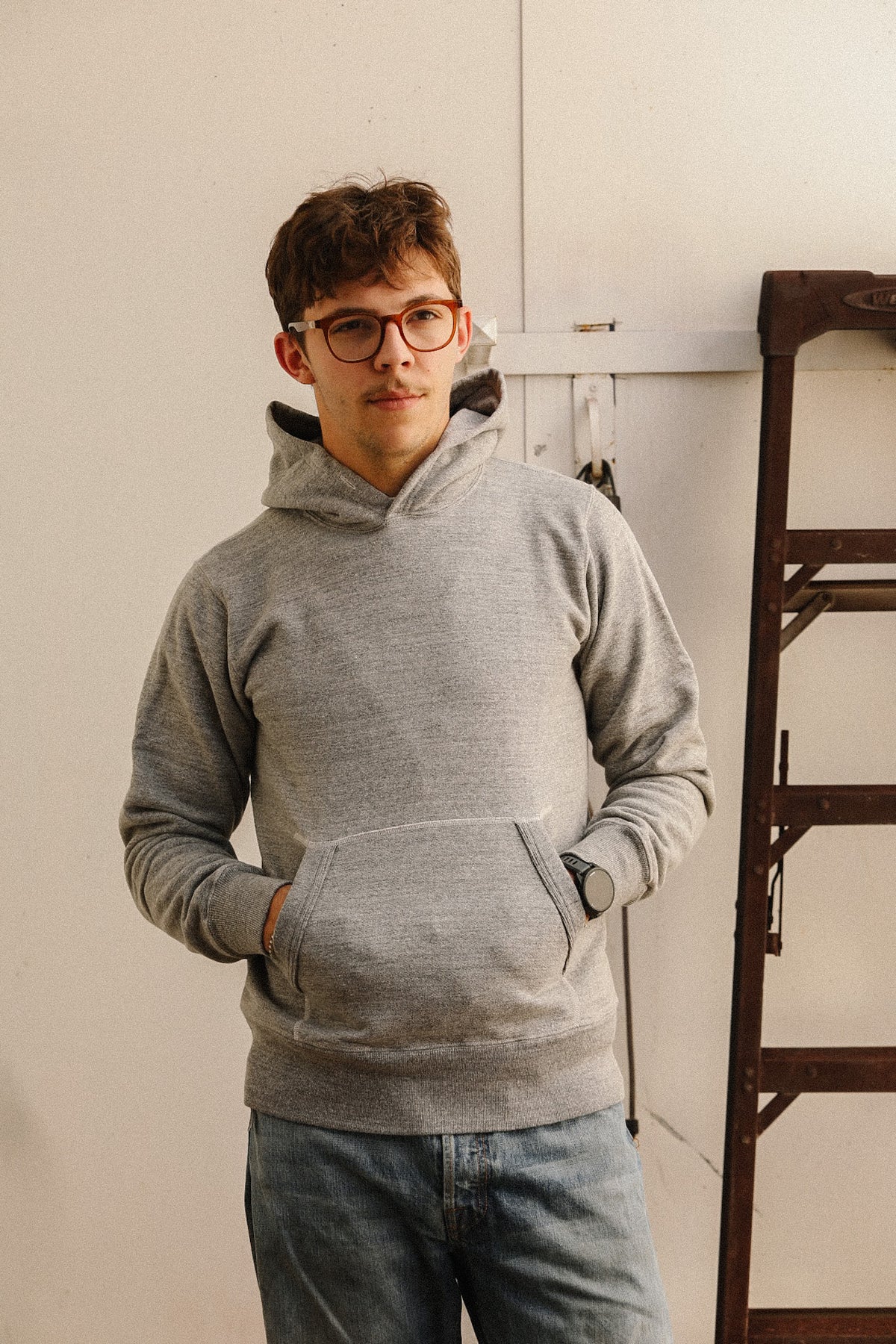 A man wearing a light gray heathered pullover hoodie with a front kangaroo pocket and glasses, standing in a room with a white wall and a wooden ladder on the right.