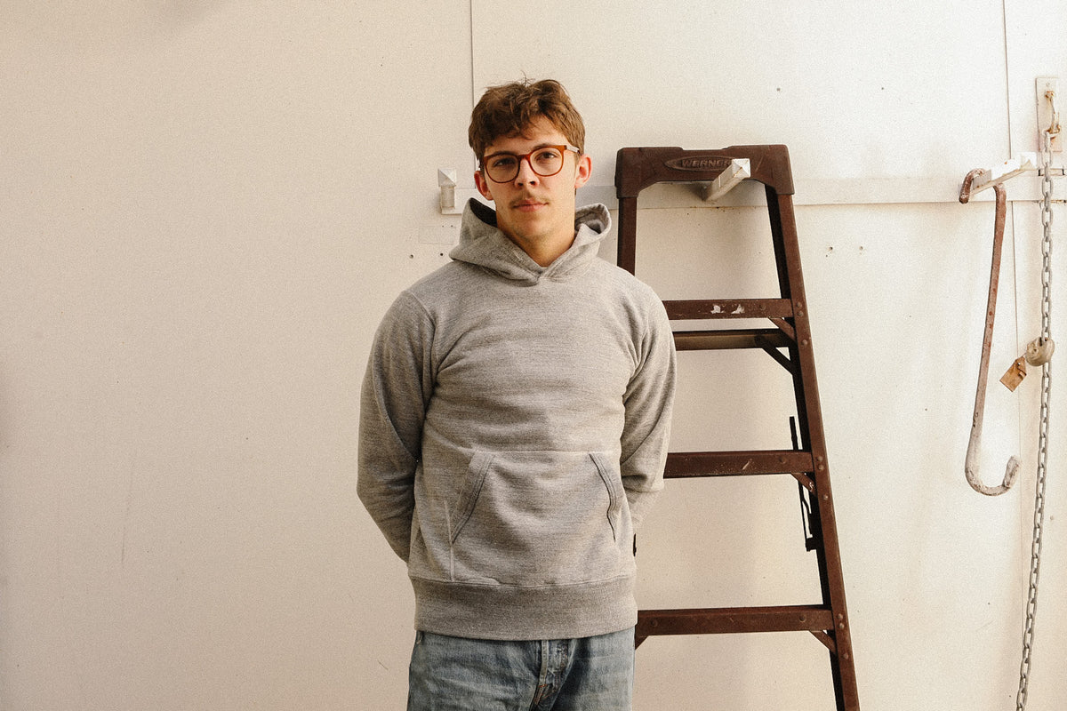 A young man with glasses stands wearing a light gray heathered hoodie and blue jeans, posing in front of a white wall with a wooden ladder behind him.
