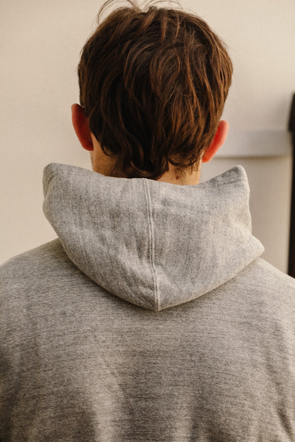 A person viewed from behind wearing a light gray heathered hoodie, showing the back of the neck, hair, and shoulders against a plain indoor wall.