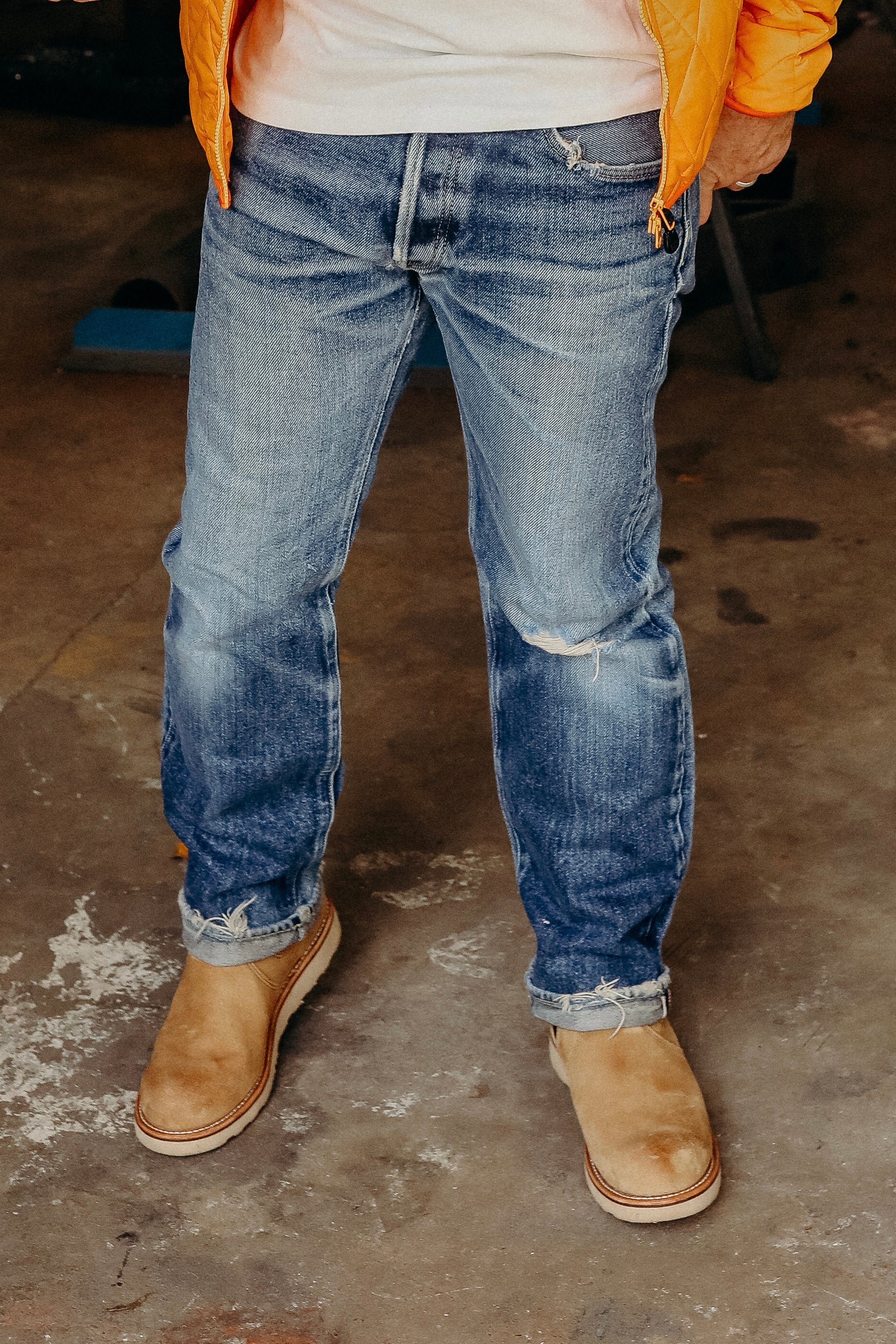 Man wearing faded blue jeans with rolled cuffs and tan suede boots standing on a concrete floor