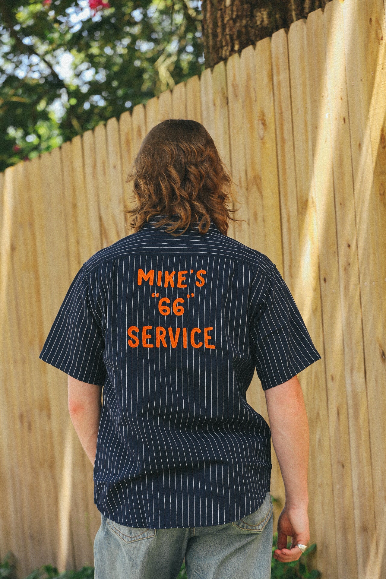 Man wearing navy blue pinstripe short sleeve work shirt with orange embroidered text 'Mike's 66 Service' on back outdoors by wooden fence