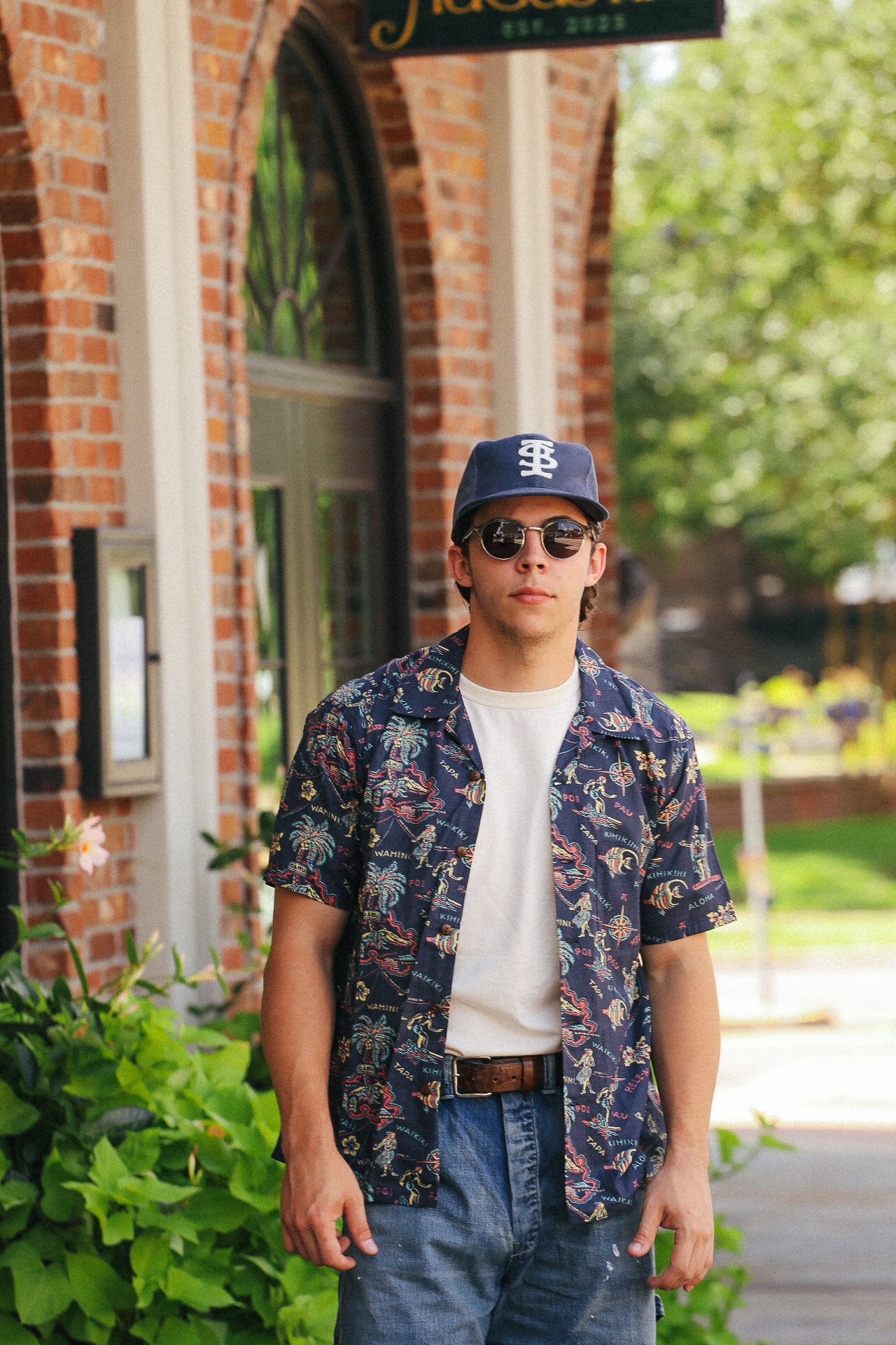 Man wearing navy cotton linen slub yarn Hawaiian shirt Hele mai e over white tee with baseball cap and sunglasses in urban outdoor setting
