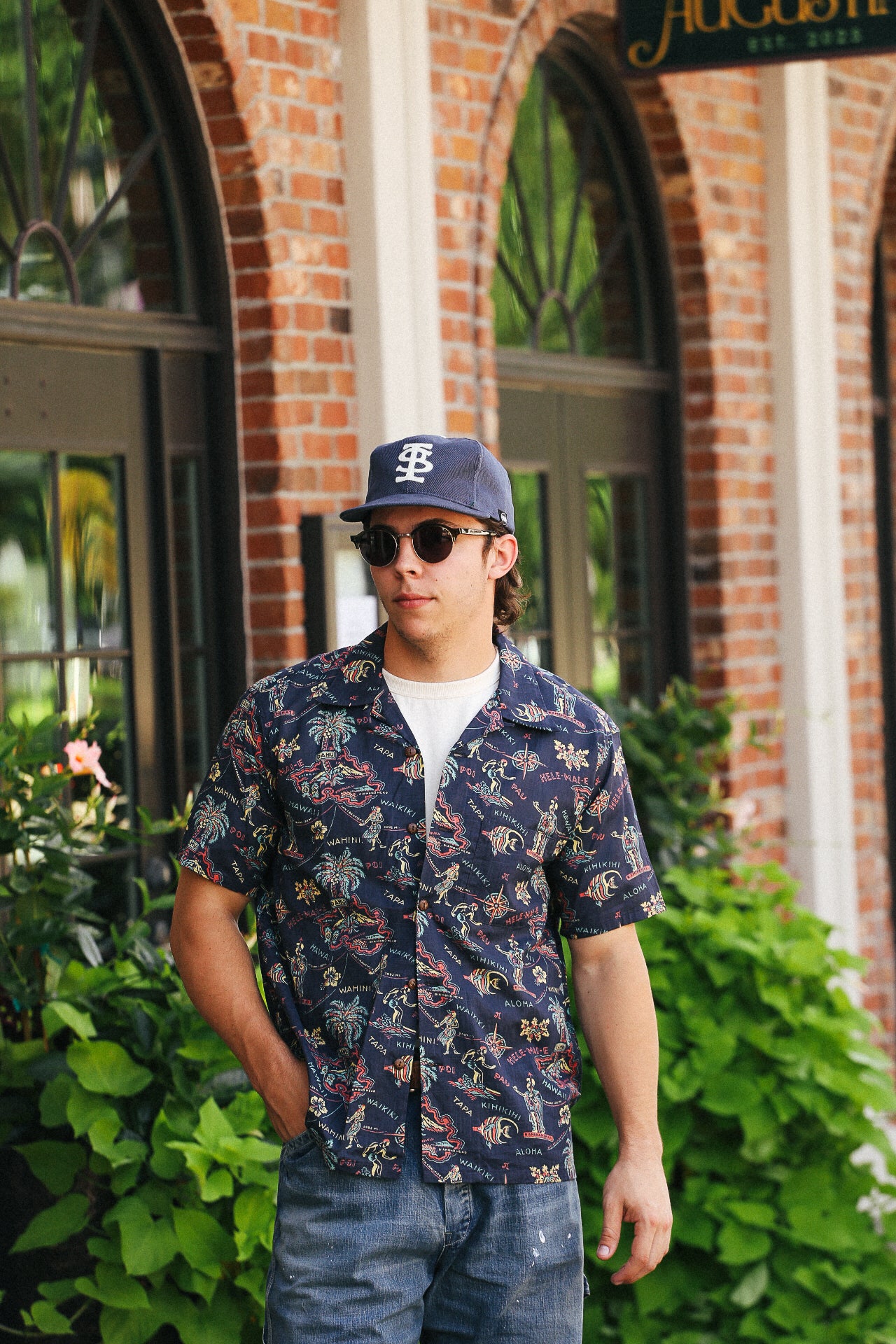 Man wearing navy Hawaiian shirt with tropical print, sunglasses, and navy cap in outdoor urban setting
