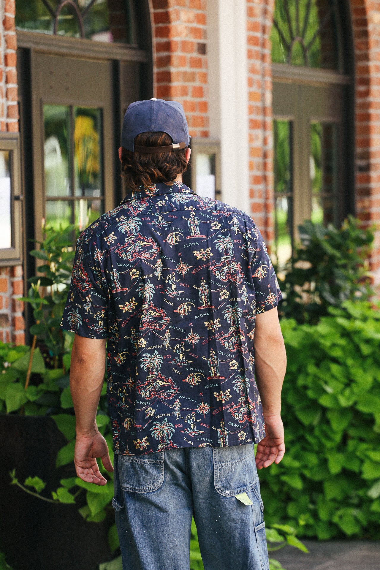 Man wearing navy Hawaiian shirt with vintage map and tropical print, blue cap, standing outside brick building