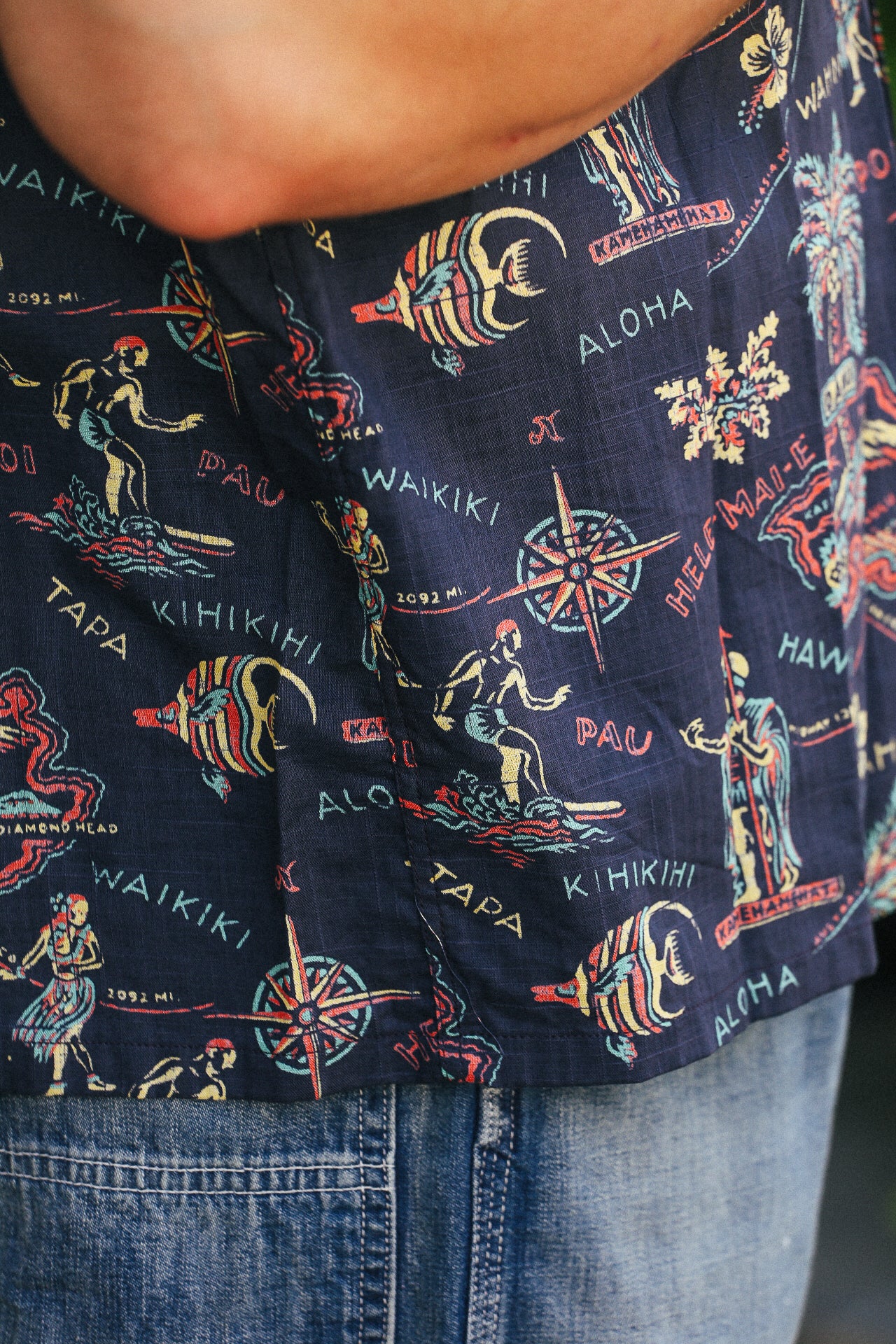 Close-up of navy blue cotton linen Hawaiian shirt with colorful island motifs and text, worn with blue jeans