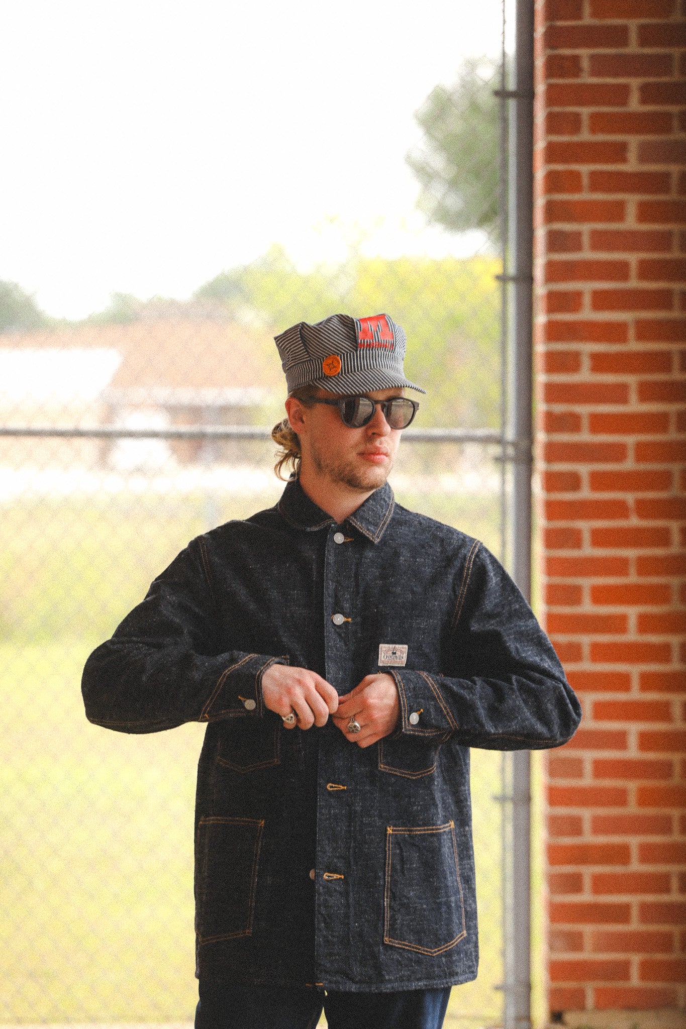 Man wearing a dark denim jacket and cap with a brick building in the background