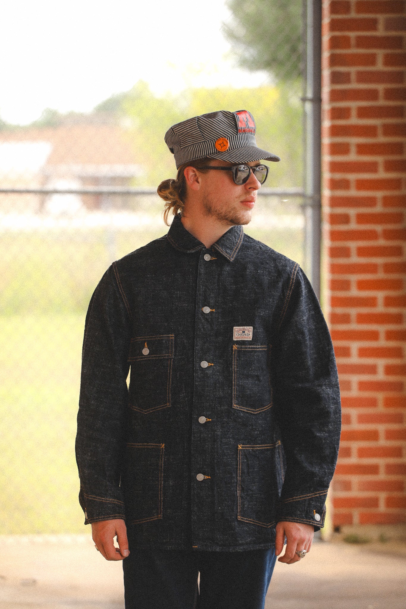 Man wearing a dark denim jacket and cap with a brick building in the background
