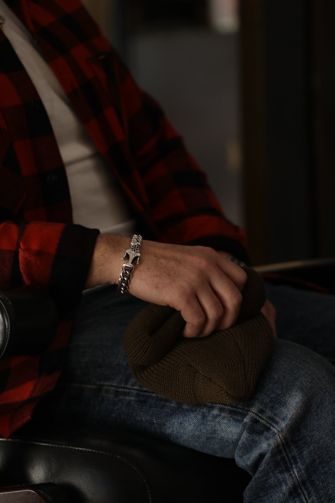 Close-up of a man wearing a silver chain bracelet with a detailed clasp, holding a folded olive green knit hat, dressed in a red and black plaid shirt and blue jeans