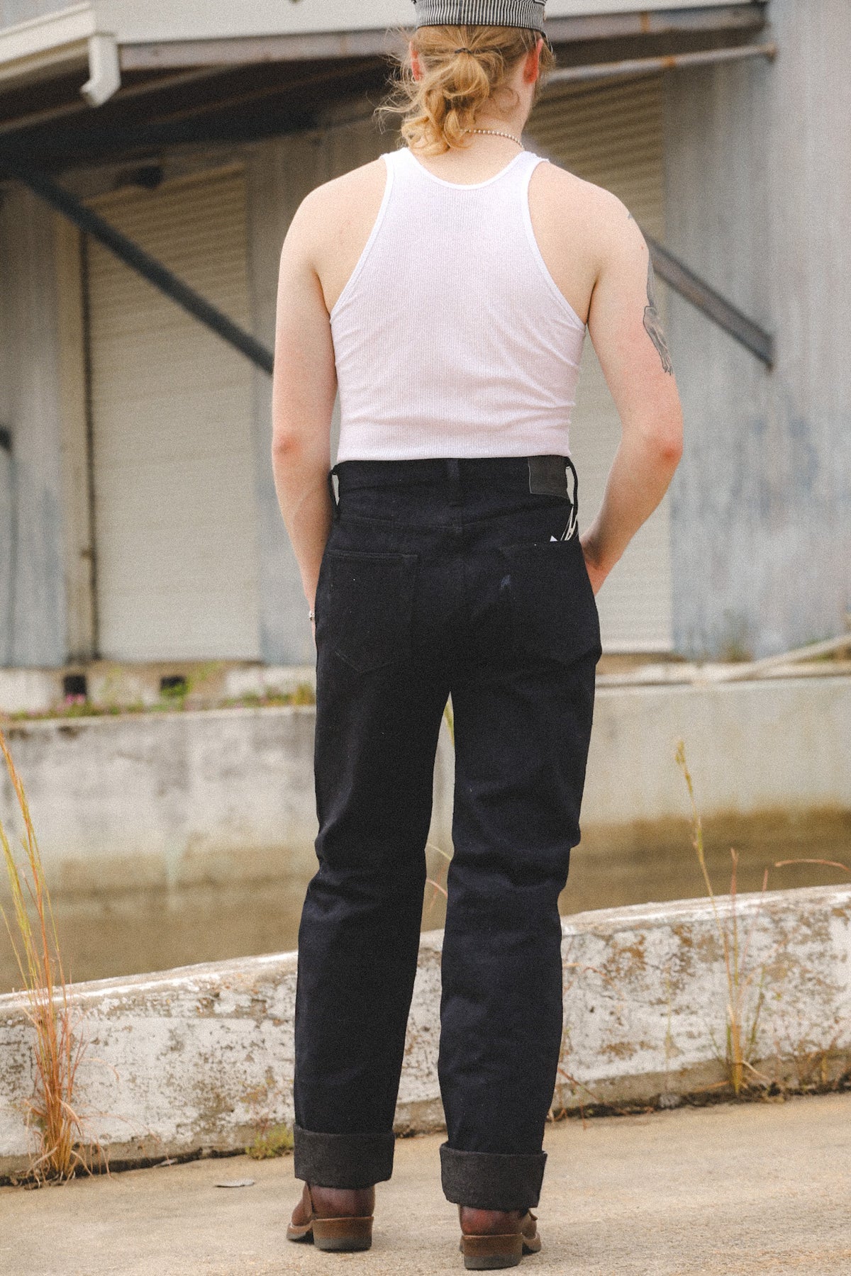 A person standing outdoors with their back to the camera, wearing dark indigo denim jeans, a white tank top, and a gray cap.