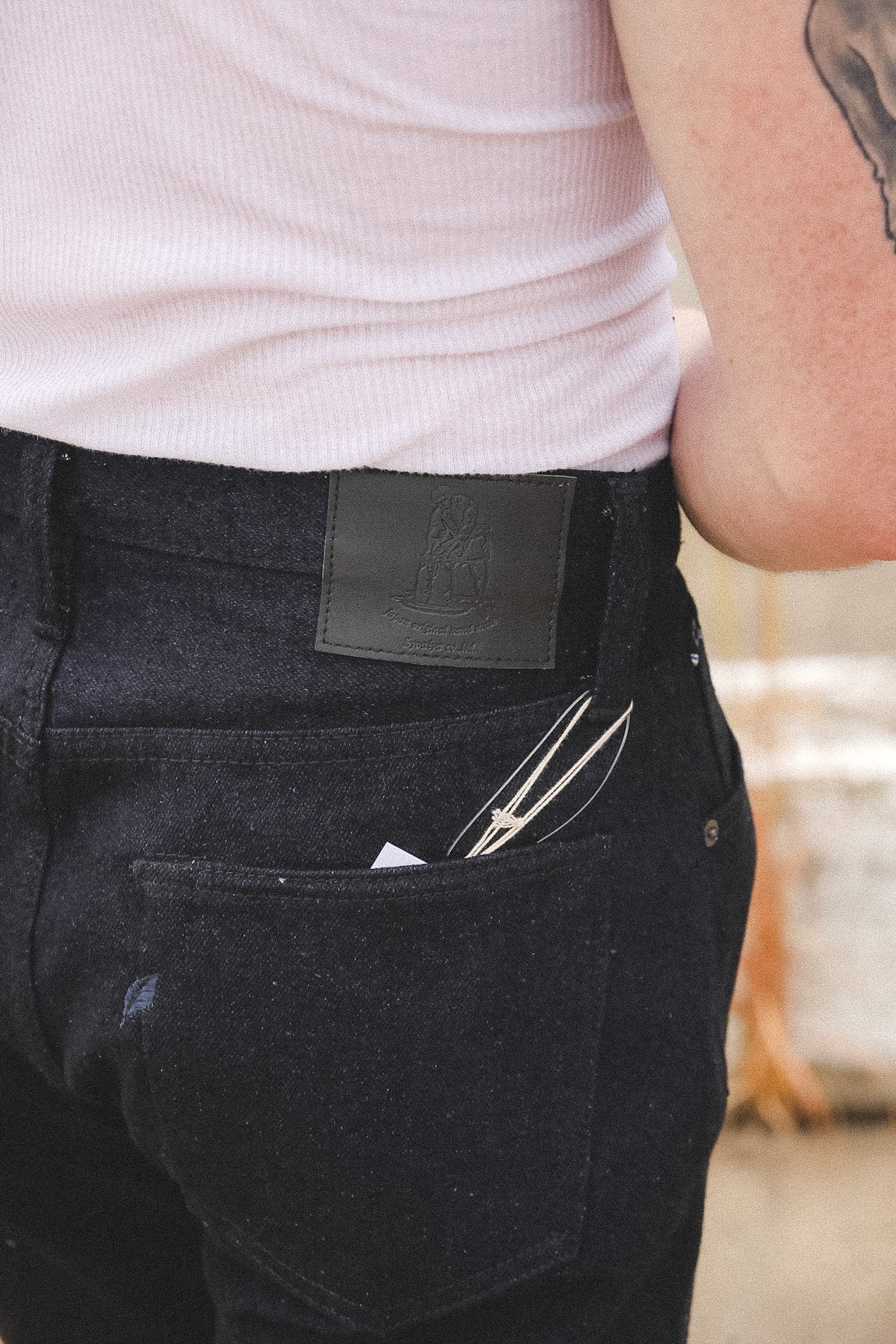 Close-up of a person wearing dark indigo denim jeans with a black leather patch on the back waistband and a back pocket holding a pair of scissors, paired with a light pink ribbed top.