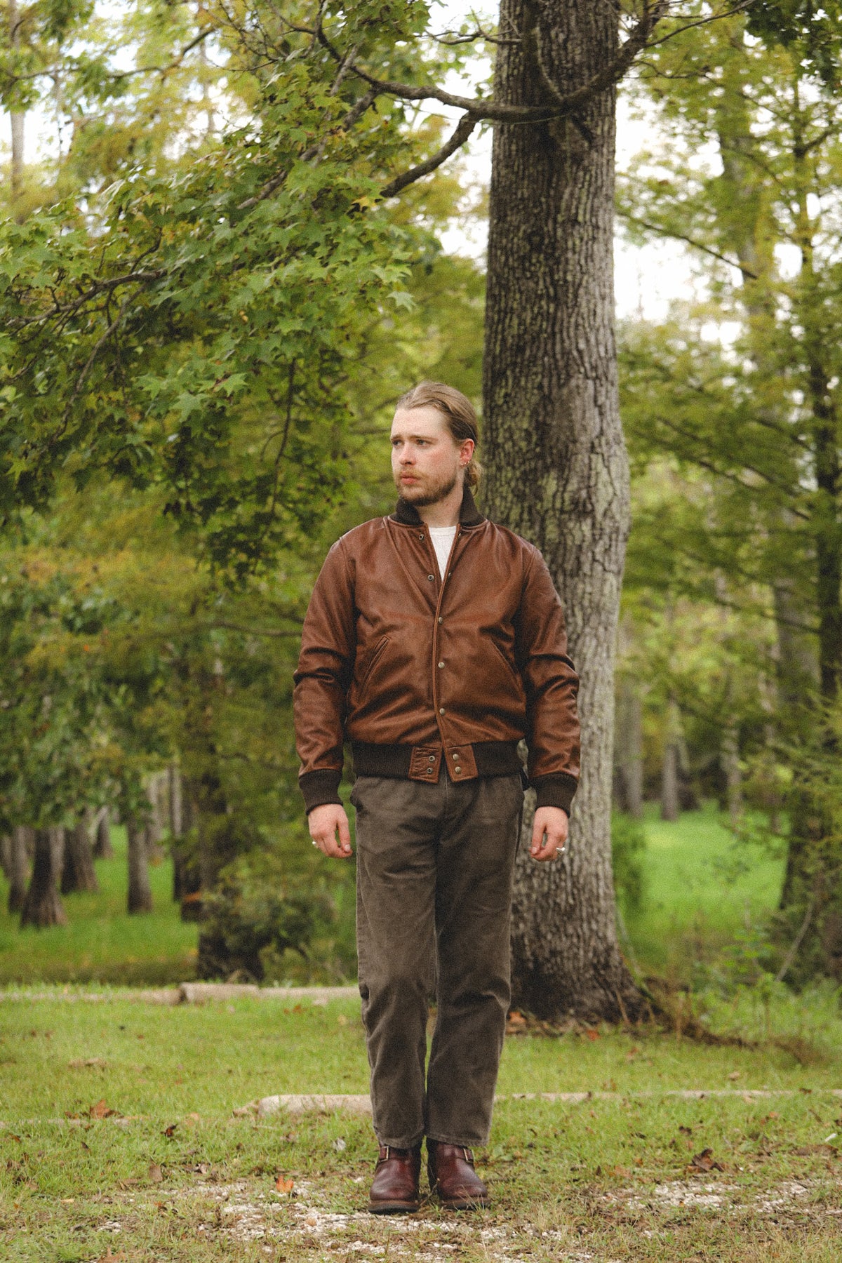 A man wearing a brown leather bomber jacket, brown pants, and brown boots stands in a grassy wooded area next to a large tree trunk.