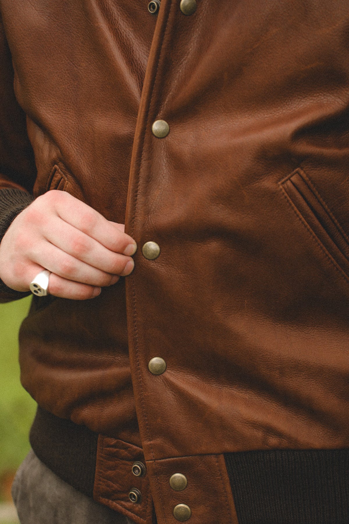 Close-up of a person wearing a brown leather jacket, with a hand pulling a metal snap button on the front. The jacket has a warm brown color, a dark ribbed collar, and brass-toned snap buttons.