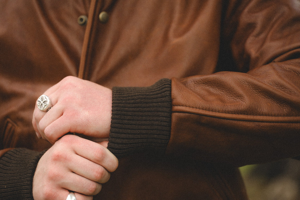 A close-up shot of a person wearing a brown leather jacket with a dark ribbed knit cuff, showing their hands and a silver-toned ring.