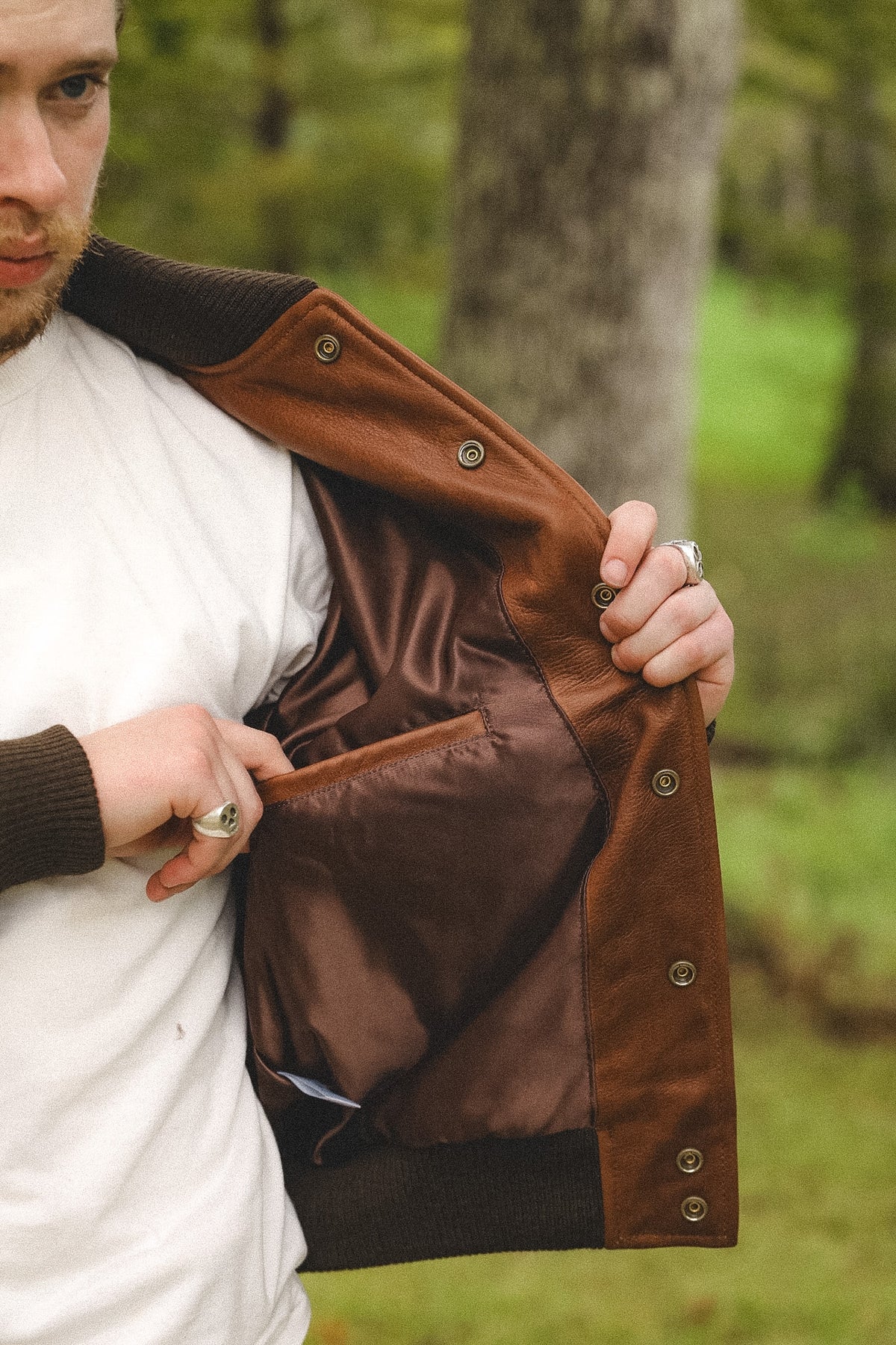 A man outdoors wearing a brown suede jacket with a brown satin-lined interior and dark ribbed knit trim, opening the jacket to show the lining.
