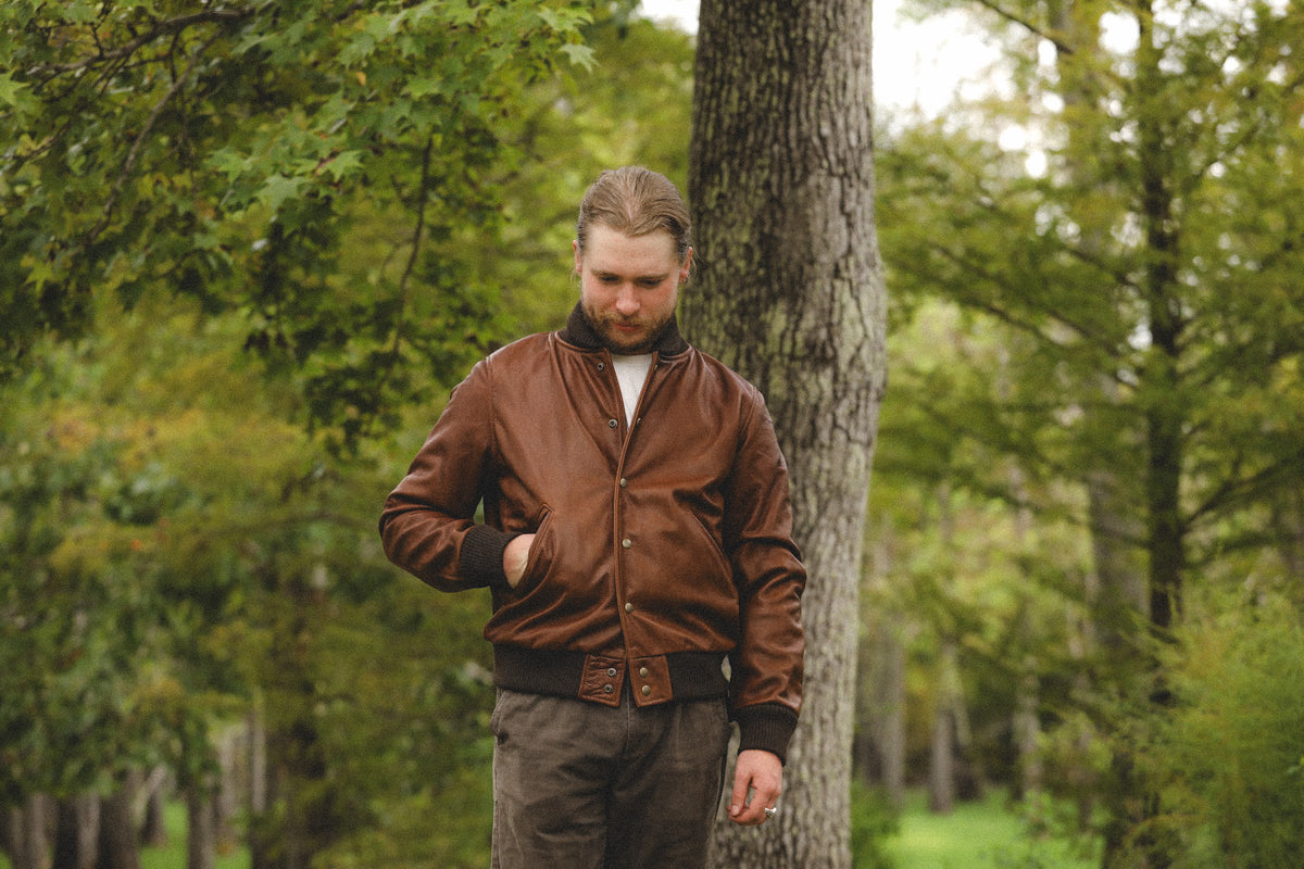 A man with a beard and long hair stands outdoors in a wooded area next to a tree, wearing a brown leather bomber jacket over a white shirt and dark brown pants.