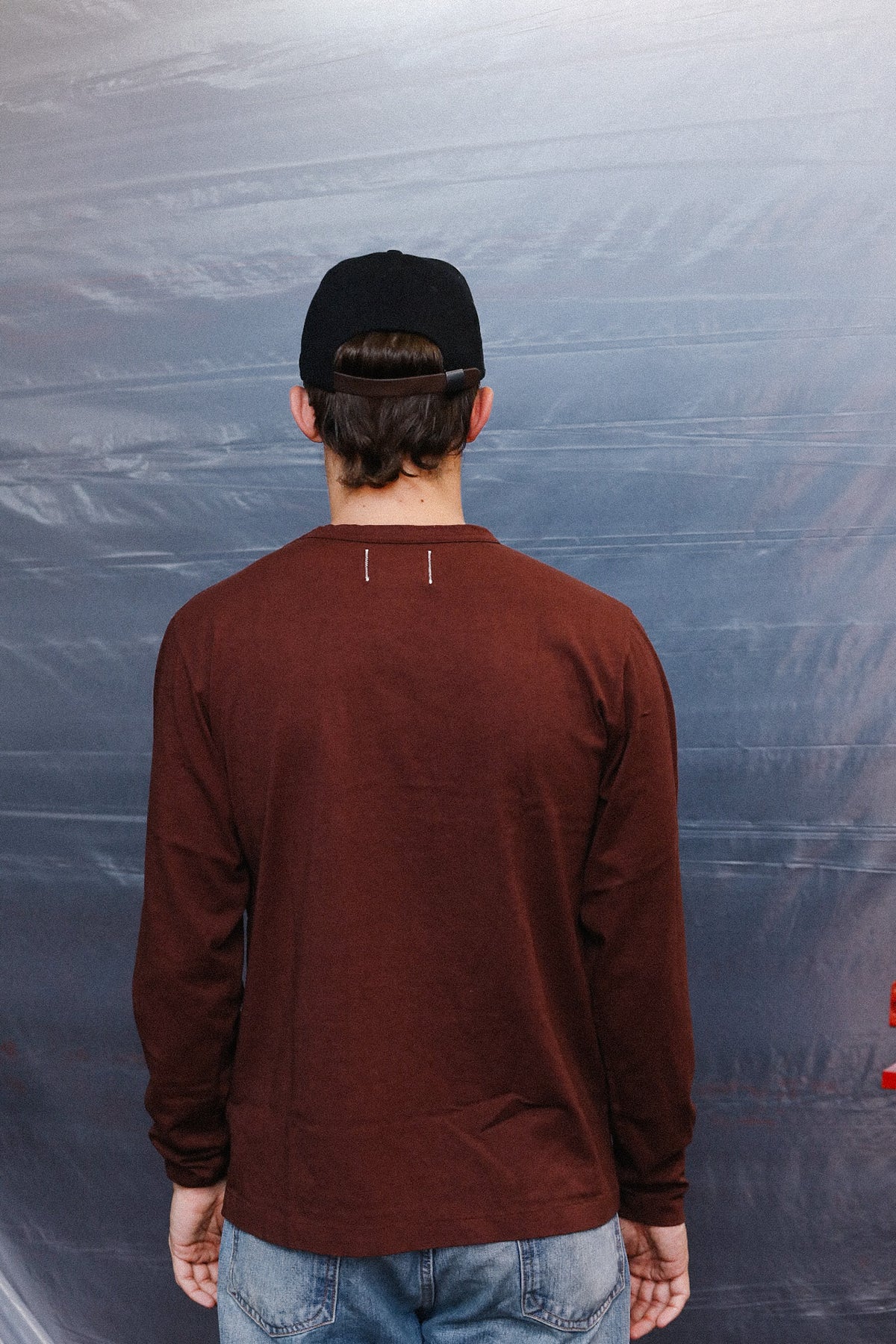 A man viewed from behind wearing a dark brown long-sleeve crewneck shirt, a black baseball cap, and blue jeans, standing against a wrinkled gray backdrop.