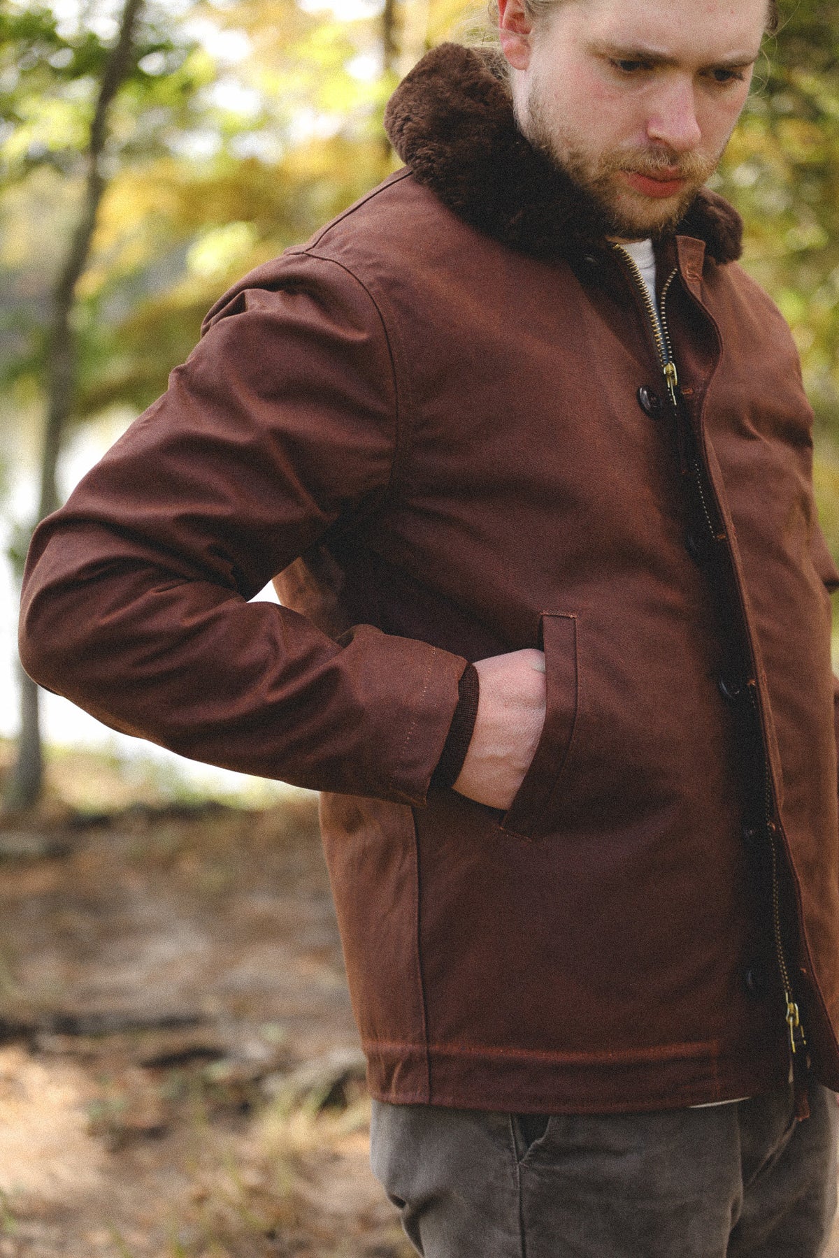 A man wearing a brown waxed canvas utility jacket with a dark brown faux-fur collar, standing in an outdoor wooded setting.