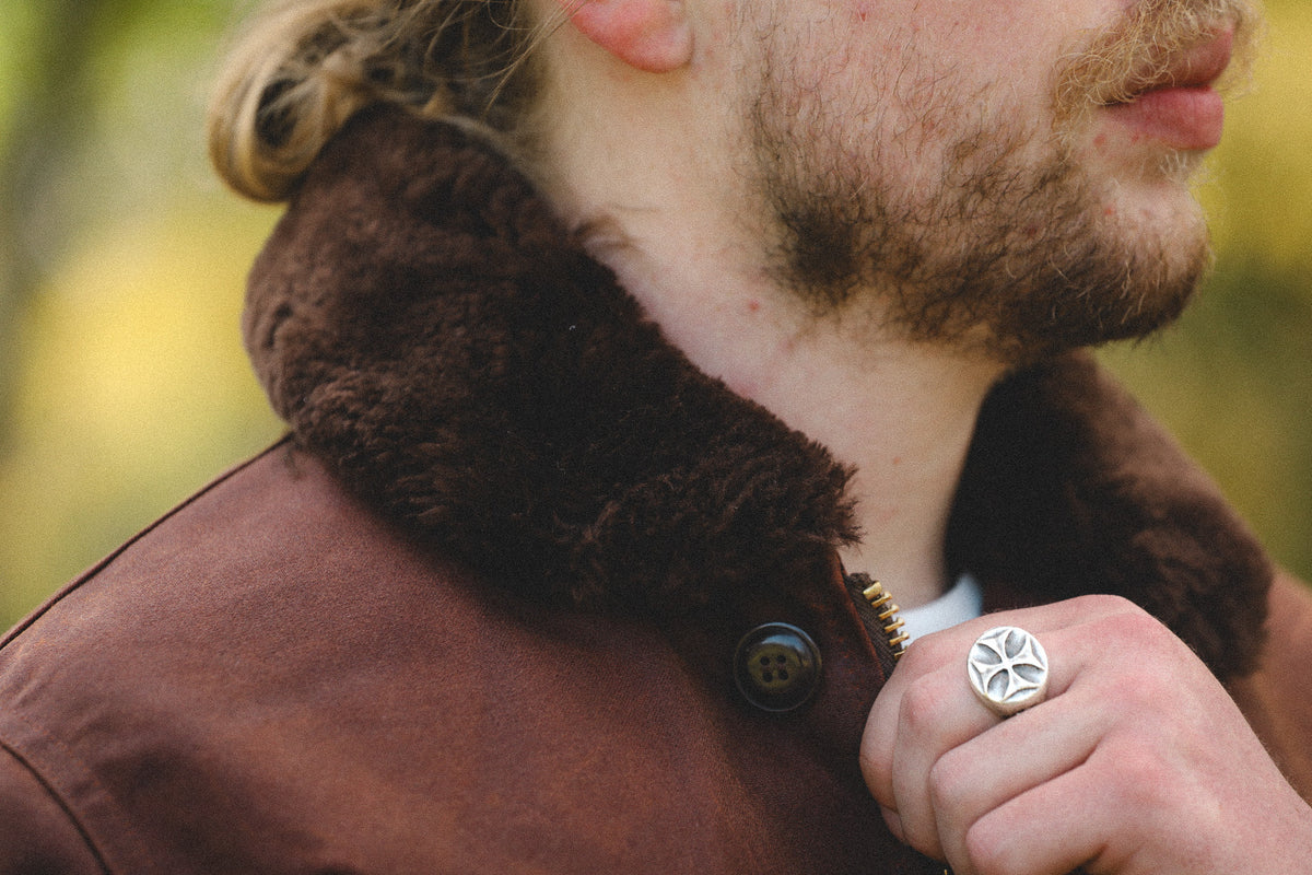 Close-up of a man wearing a brown jacket with a dark brown faux-shearling collar and a silver-toned ring on his finger, set against a blurred outdoor background.