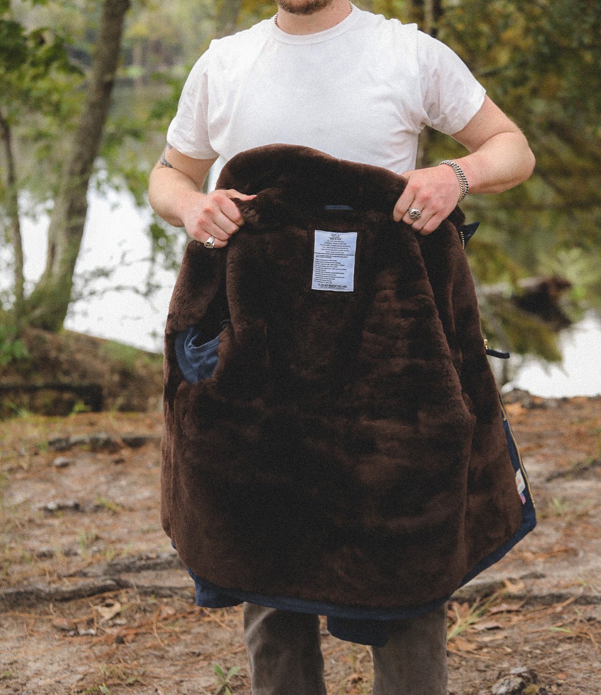 A man outdoors by a lake holds up a dark brown plush fleece-lined jacket or coat, showing the soft interior lining and a small sewn-in label.