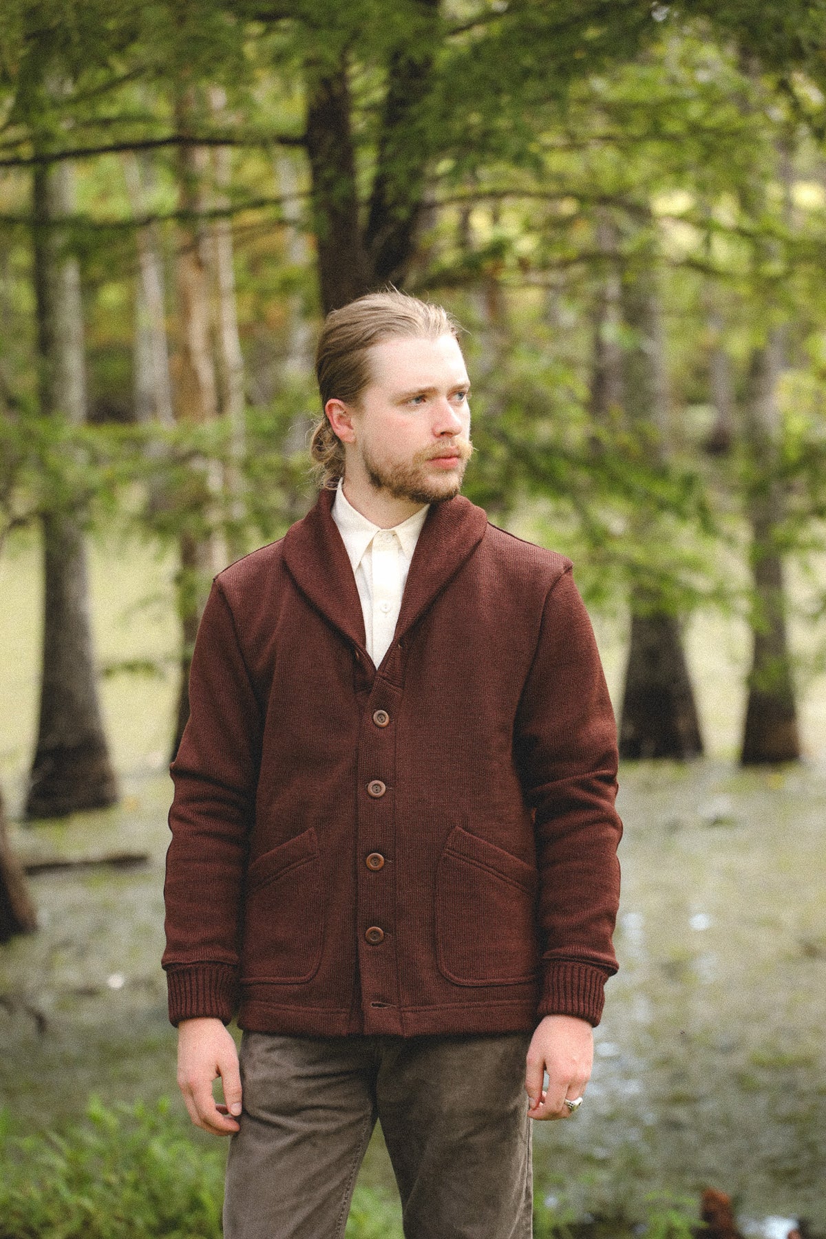 A man wearing a dark brown shawl-collar button-front cardigan over a white collared shirt, standing in a wooded outdoor setting.