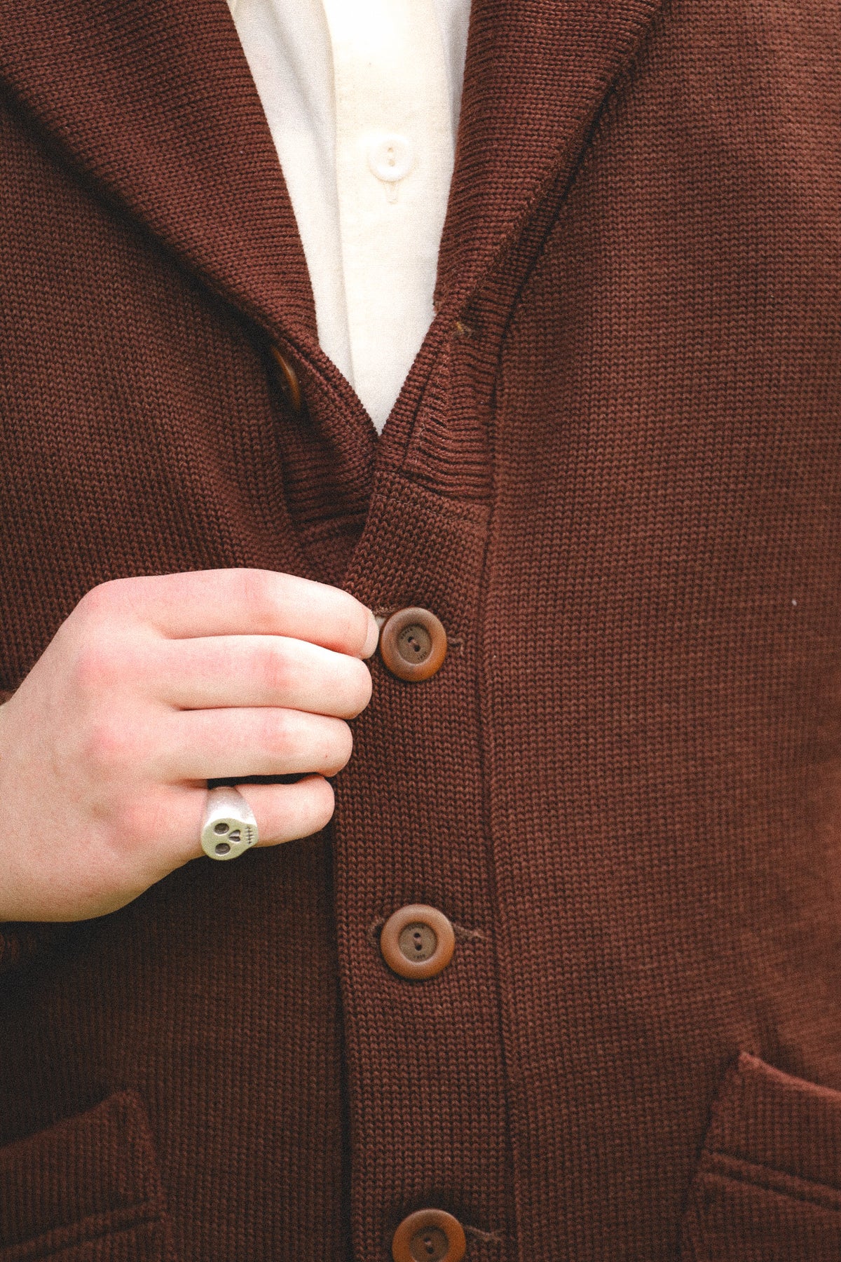 A close-up of a person wearing a brown textured knit cardigan over a cream button-up shirt, with a hand adjusting the front buttons.