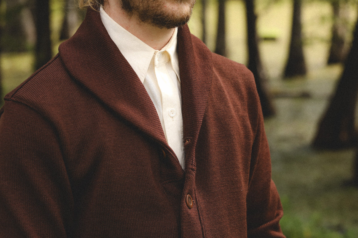 A close-up of a man wearing a dark brown textured knit cardigan over a cream button-up shirt, standing in a blurred forest setting.