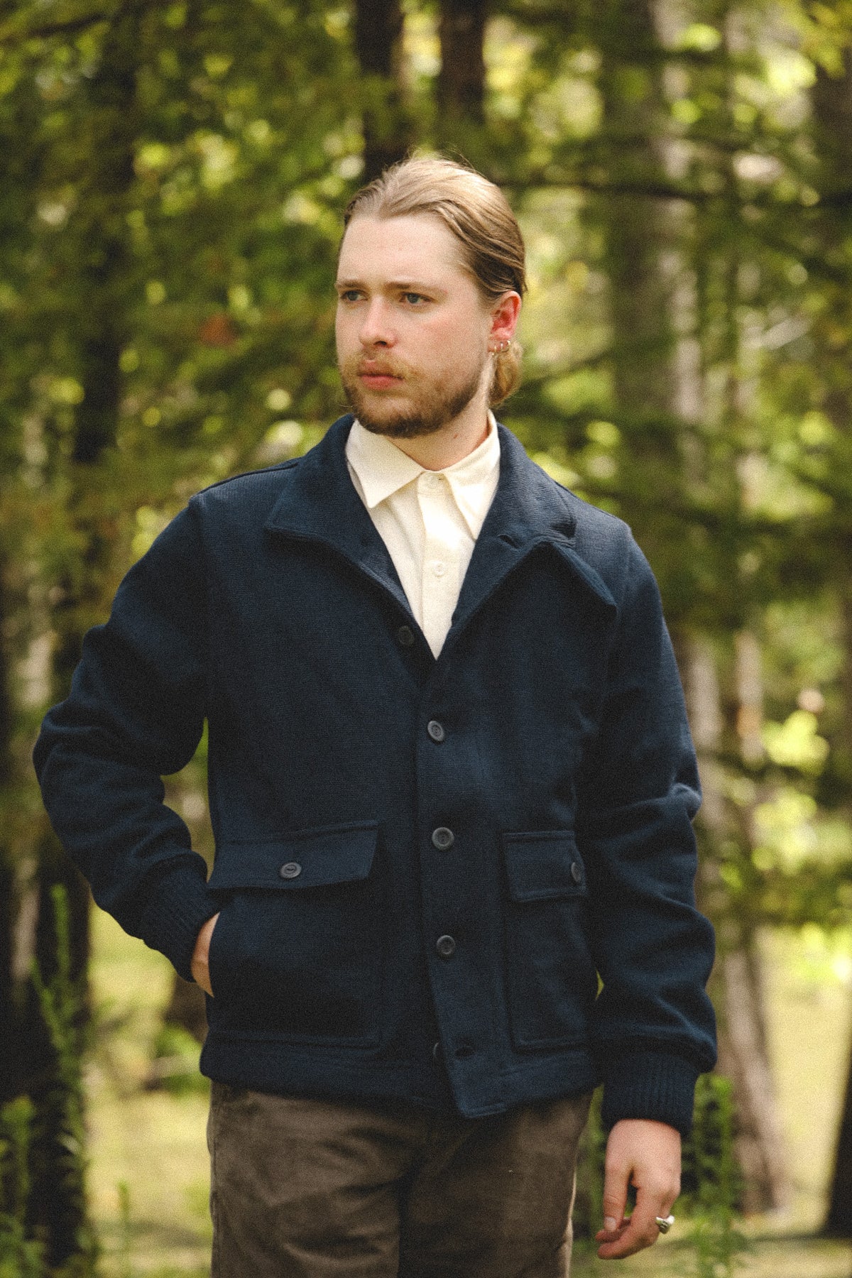A man wearing a dark navy blue corduroy jacket over a cream shirt and brown trousers, standing in a sunlit forest.