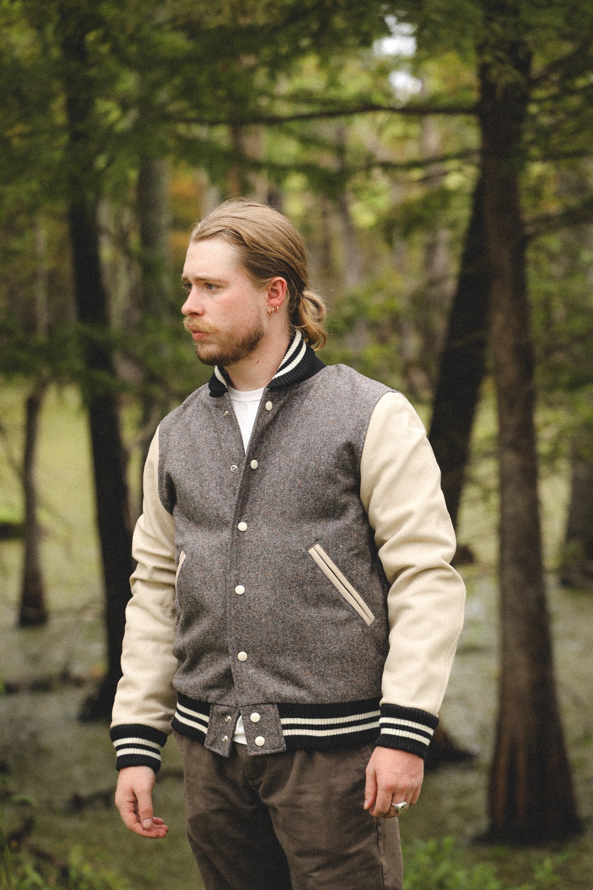 A man wearing a gray and cream varsity jacket stands in a wooded outdoor setting, looking to the side.