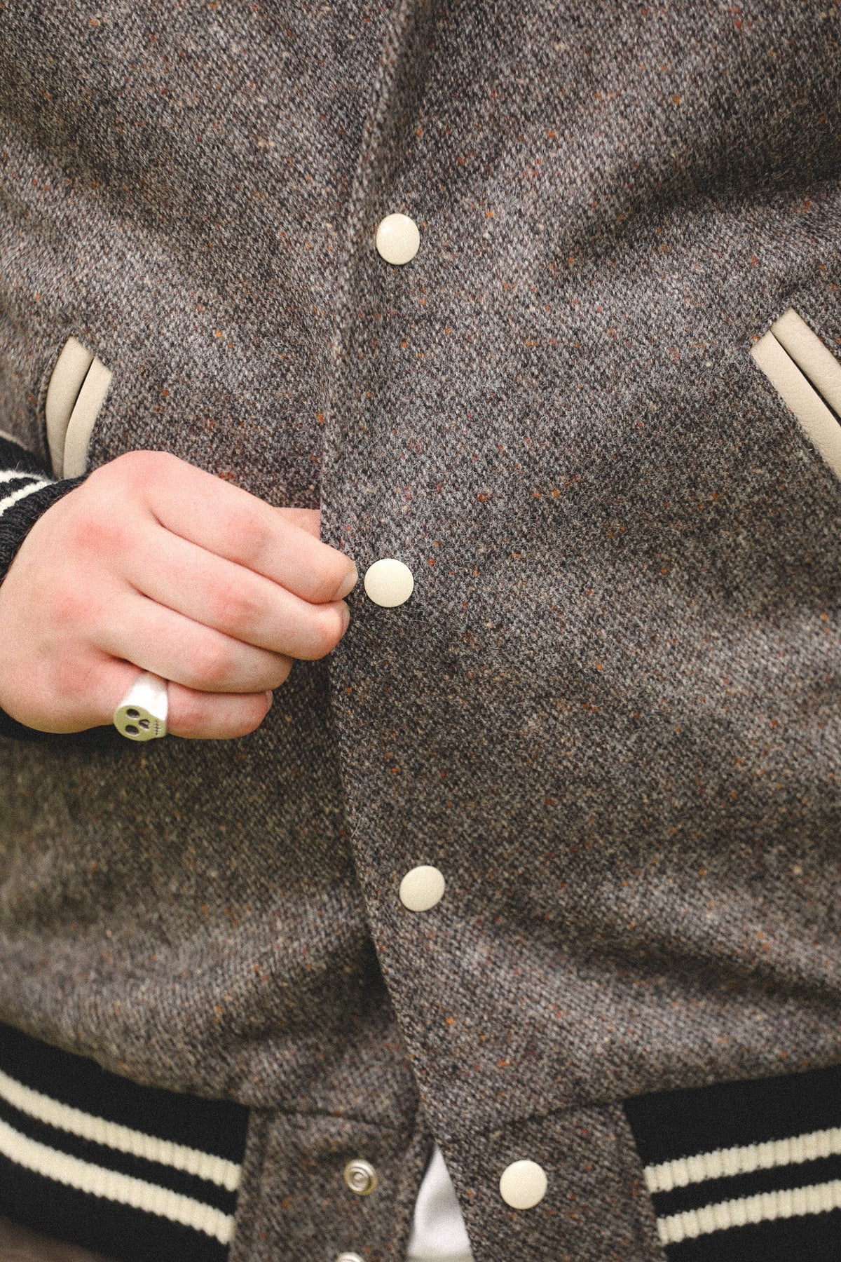 Close-up of a person wearing a brown tweed varsity jacket, showing the front buttons, ribbed knit collar and cuffs with white stripes, and a hand adjusting the collar.
