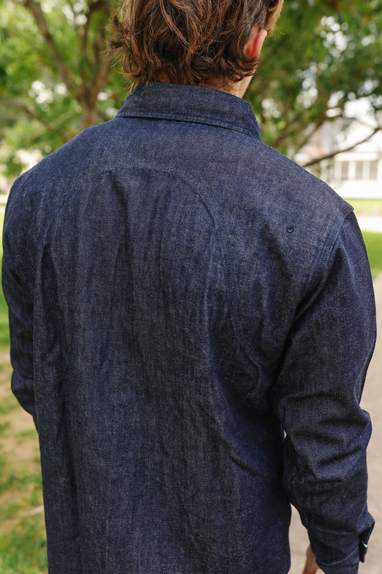 Back view of person wearing indigo denim medical shirt outdoors with greenery and house background