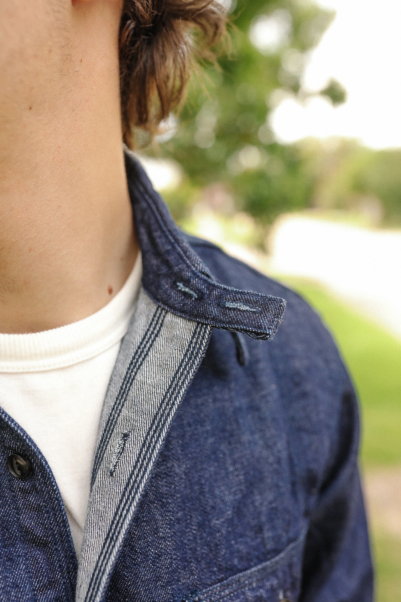 Close-up of a person wearing an indigo denim medical shirt with visible collar and buttonhole details over a white crewneck shirt outdoors