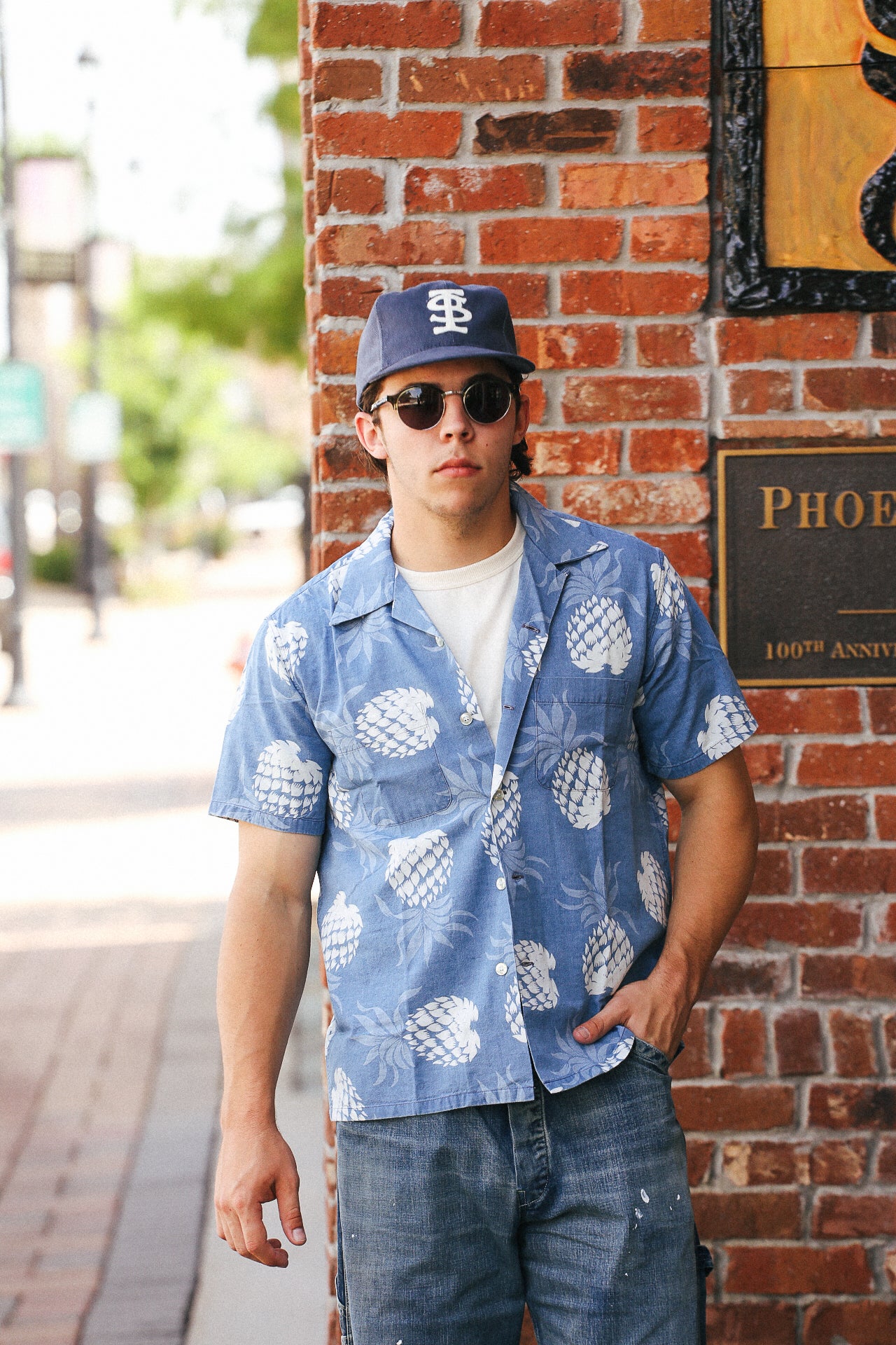 Man wearing Duke Kahanamoku indigo cotton Hawaiian shirt with white pineapple print, sunglasses, and baseball cap leaning against red brick wall